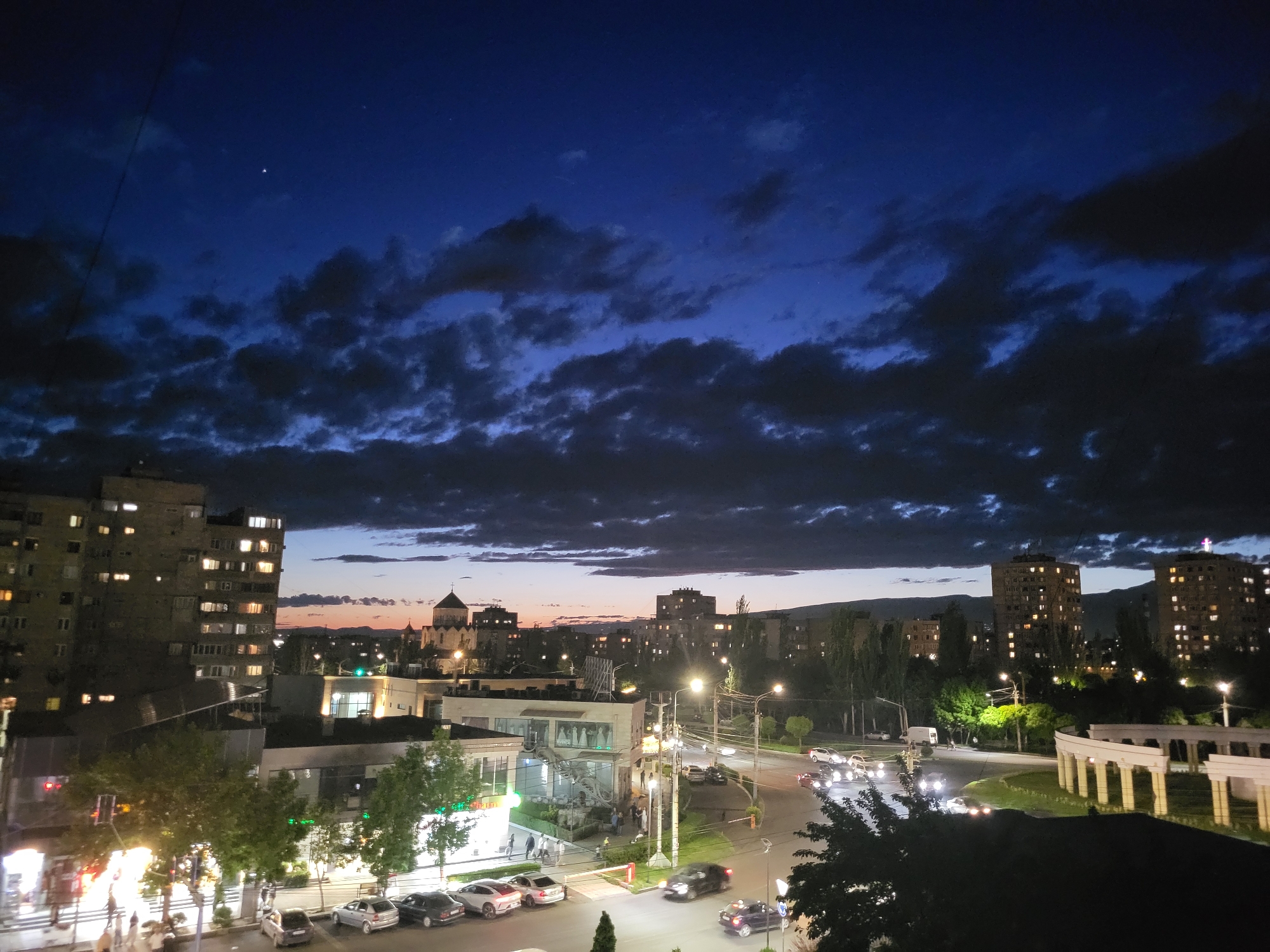 The image depicts a night view of Yerevan, Armenia, characterized by a cityscape under a cloudy, dark blue sky. A single visible star punctuates the upper left portion of the sky. Buildings of varying heights, some illuminated with multiple lit windows, define the urban landscape. A religious structure with a dome and cross atop is visible amidst the buildings. The streets are lit by streetlights, and cars are seen moving along the roadways. A pedestrian crossing is visible, and several parked cars line the streets. Trees punctuate the cityscape, adding patches of green to the urban environment. The overall scene presents a blend of residential and commercial areas with active nighttime traffic.