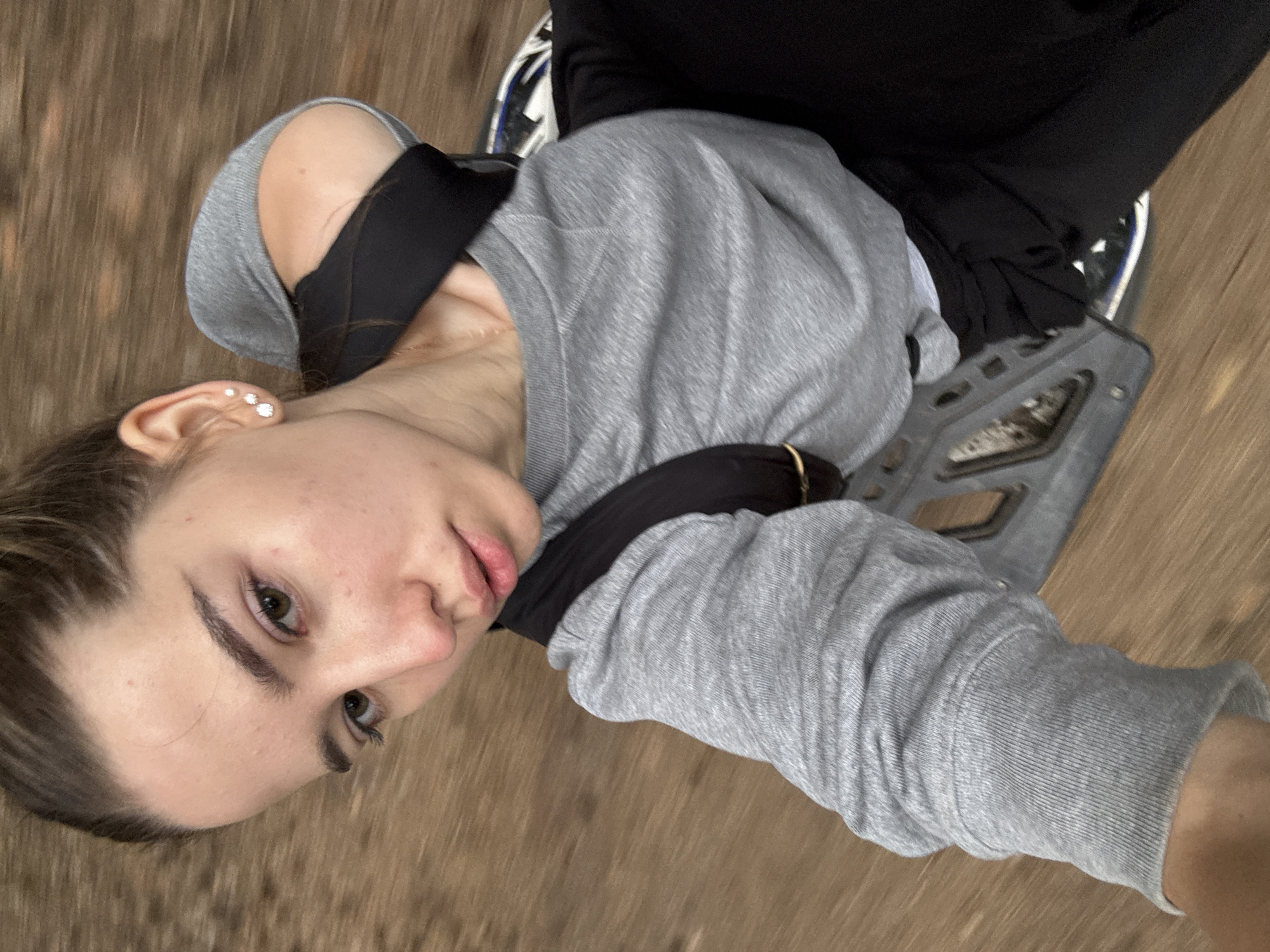 A young woman with her hair pulled back in a bun is taking a selfie. She is wearing a grey sweatshirt and black pants. She has a backpack on. The background is blurry and appears to be a wooden floor or surface. The image was taken in Noviy, Russia.