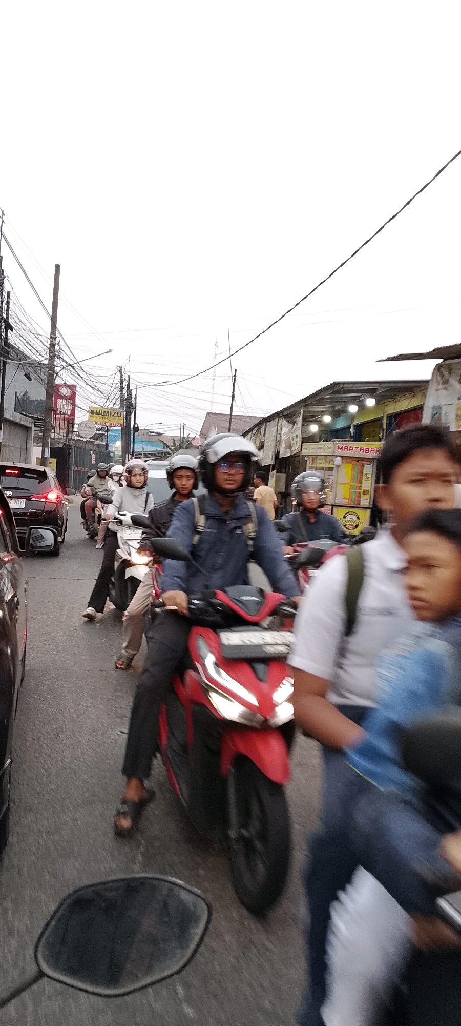 In Depok, Indonesia, a traffic jam unfolds on a cloudy day. The scene is dominated by a procession of motorcycles and a few cars navigating a moderately busy street lined with shops and utility poles laden with electrical wires. The foreground features the driver and passenger of a red motorcycle, both wearing helmets. The driver, a male, is in focus, clad in a dark blue denim jacket and dark pants, while the passenger, a young boy, is partially obscured, wearing a blue denim jacket. Behind them, other motorcycles carry riders and passengers, all also equipped with helmets. A black car is visible on the left, its taillights illuminated. The backdrop reveals the typical urban landscape of Depok, with low-rise buildings and overhead power lines. The overall impression is one of everyday city life, characterized by vehicular congestion and pedestrian activity.