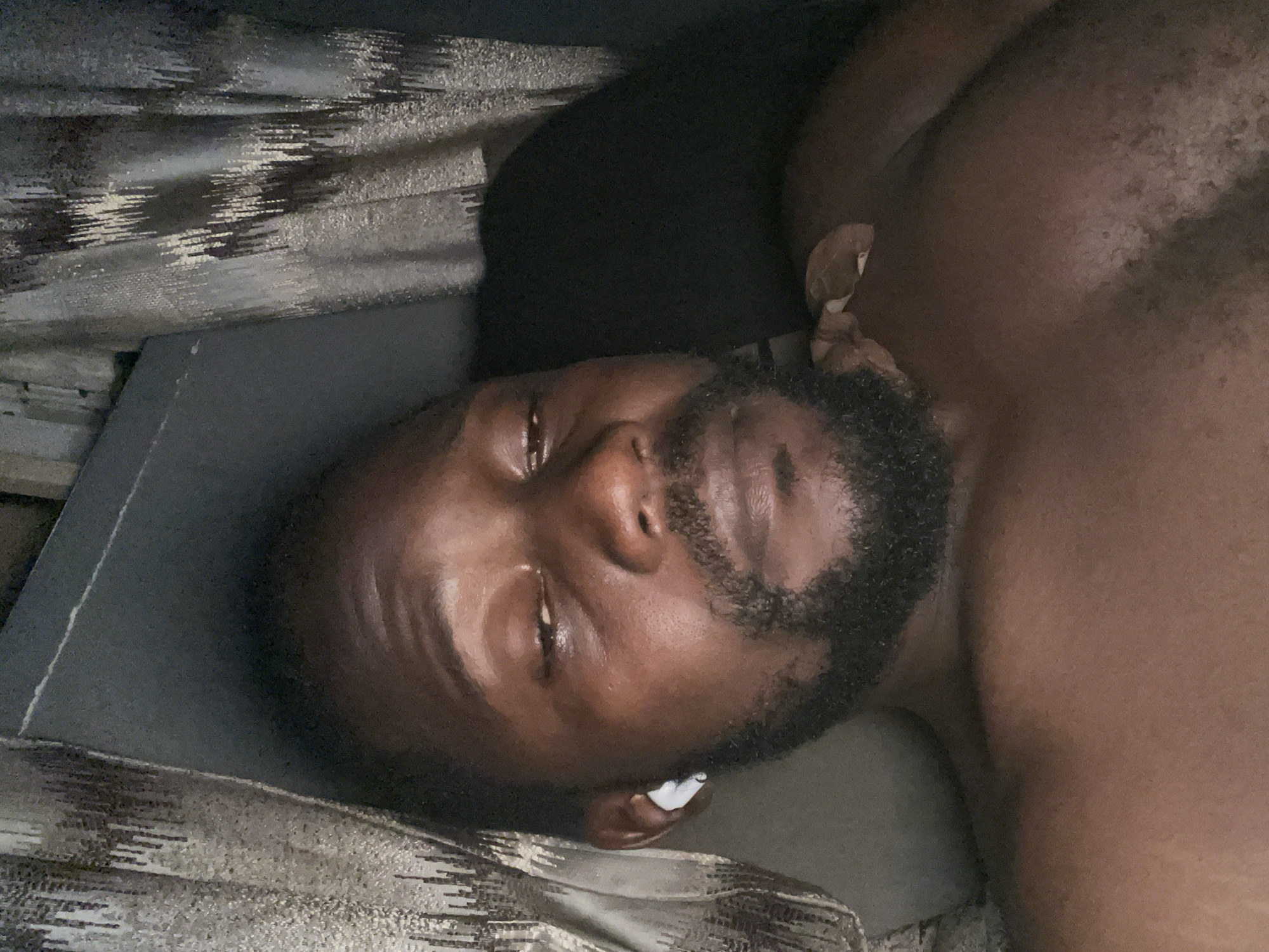 In Kurunduma, Nigeria, a medium-close-up selfie is captured, featuring a Black man lying down in what appears to be a bedroom. The man is shirtless and has dark skin and a beard. He is wearing a white earbud in his left ear. The background is dark and somewhat blurred, with hints of patterned curtains visible on either side. The lighting suggests an indoor environment, possibly at night or in a dimly lit room.