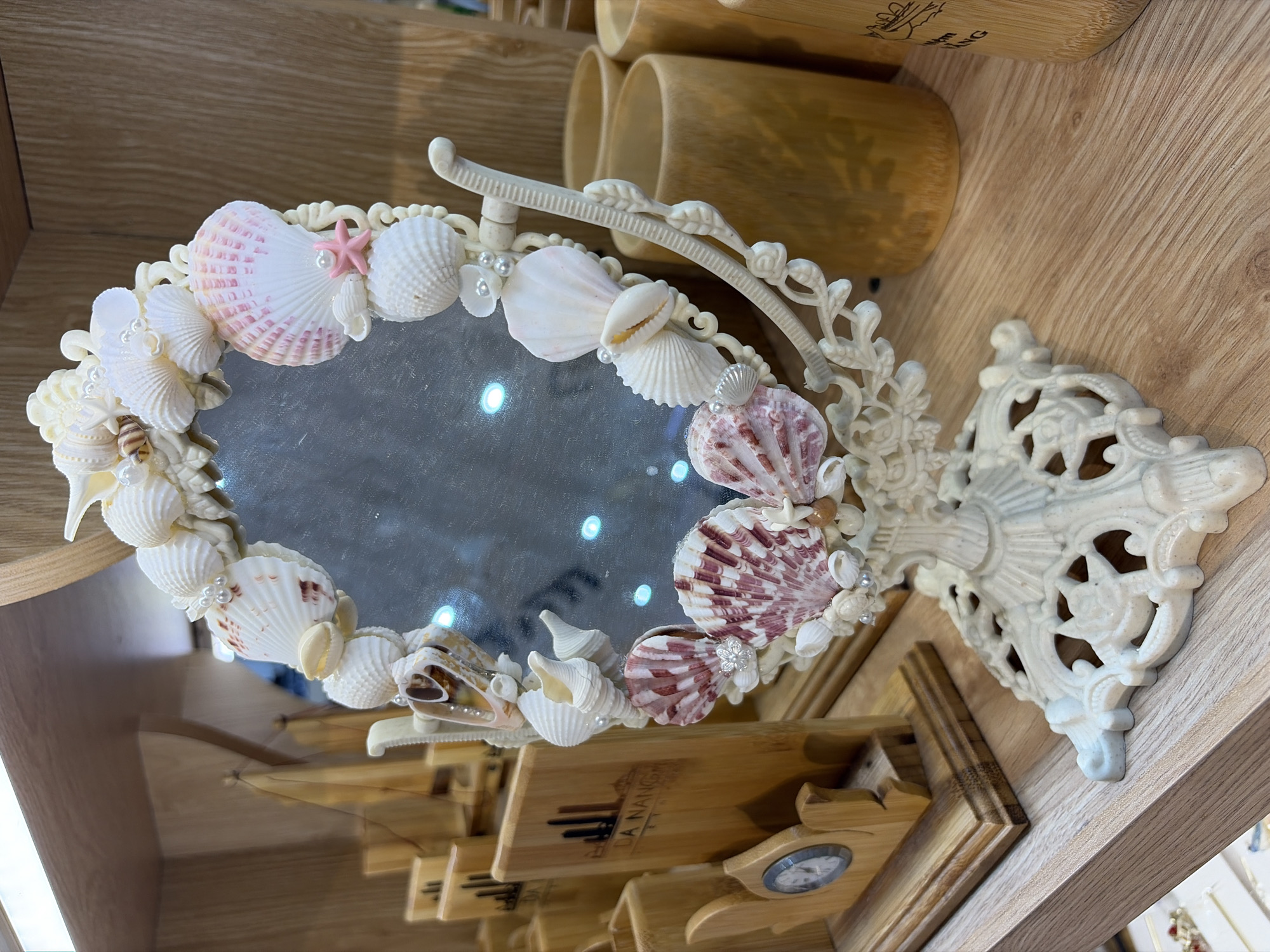 A seashell-encrusted mirror stands on a wooden shelf in what appears to be a shop in Da Nang, Vietnam. The mirror is framed with various seashells, pearls, and a small pink starfish. The stand of the mirror is ornate and cream-colored. To the left of the mirror, there are wooden display stands with what look like business cards or promotional materials, one of which has the text "DA NANG" visible. Also on the left is a small wooden clock. In the background and to the right, there are several cylindrical wooden containers. The overall impression is that of a souvenir shop display.