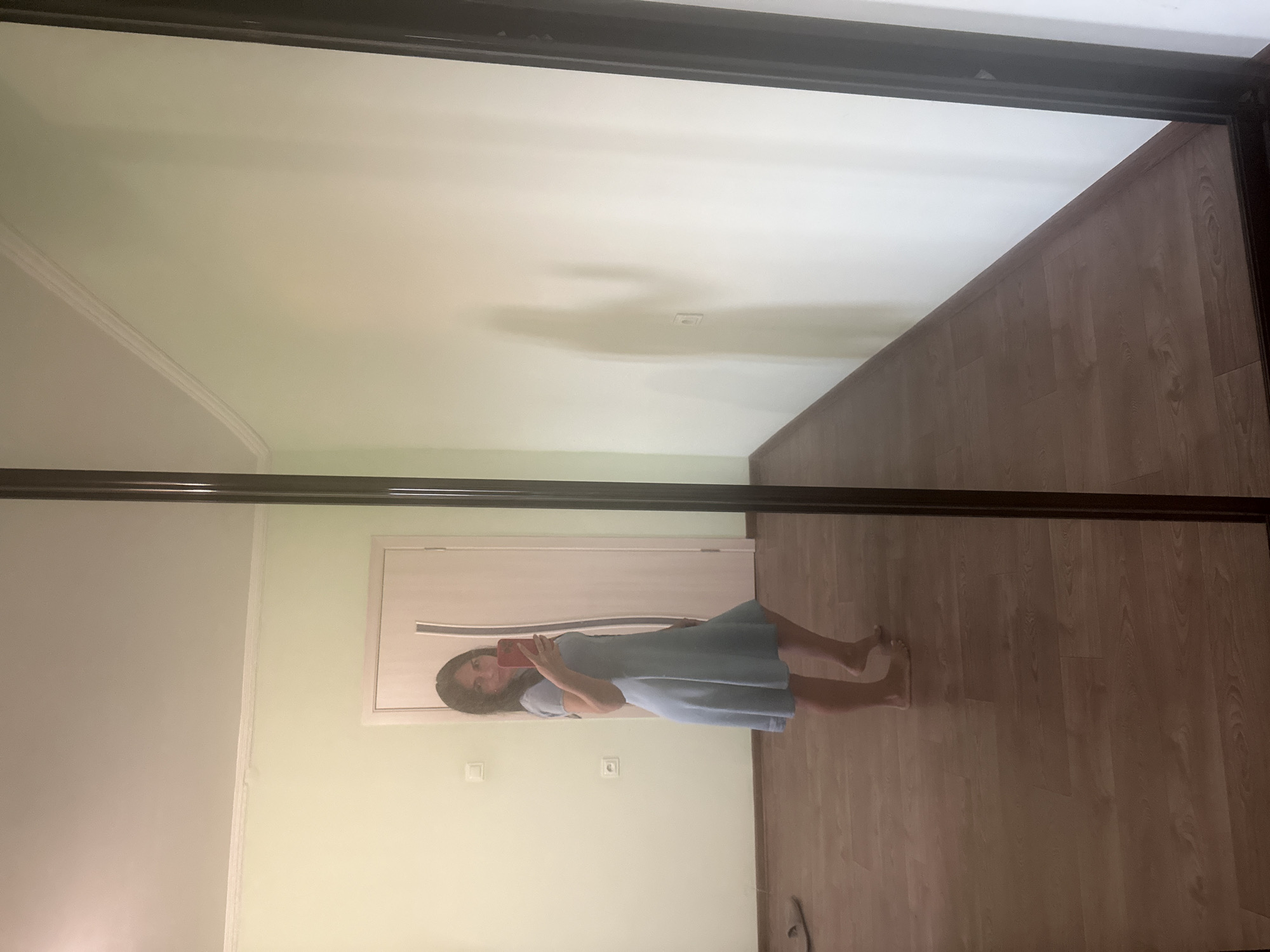 A woman, appearing to be of Eastern European descent with dark hair, stands in a room and takes a selfie in a full-length mirror. She is wearing a light blue, short-sleeved A-line dress that falls to her mid-thigh. The woman is barefoot and her right hand is raised, holding a red smartphone. She is positioned to the left of the frame, facing the mirror and slightly angled towards the viewer. Behind her, a white door with vertical dark accents is visible. The room has light green walls and a wooden floor. The mirror reflects the room, including a shadow on the wall that suggests the presence of another person or an object. The image is taken from a perspective that captures the woman and a significant portion of the room's reflection. There are two light switches on the wall. A single slipper is visible on the floor in the reflection.