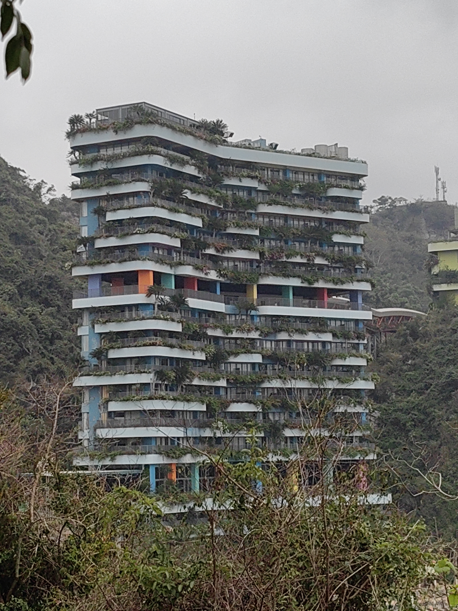 A multi-story building stands amidst dense vegetation in Hải Phòng, Vietnam. The building features cantilevered balconies on each floor, heavily populated with greenery. Painted columns in blue, orange, red, and yellow punctuate the building's façade. The structure is surrounded by lush trees and foliage. In the background, a transmission tower and adjacent building are visible on a hilltop. A pedestrian bridge is located behind the main building. In the foreground, bare branches and leaves obscure part of the lower portion of the structure. The sky is overcast.