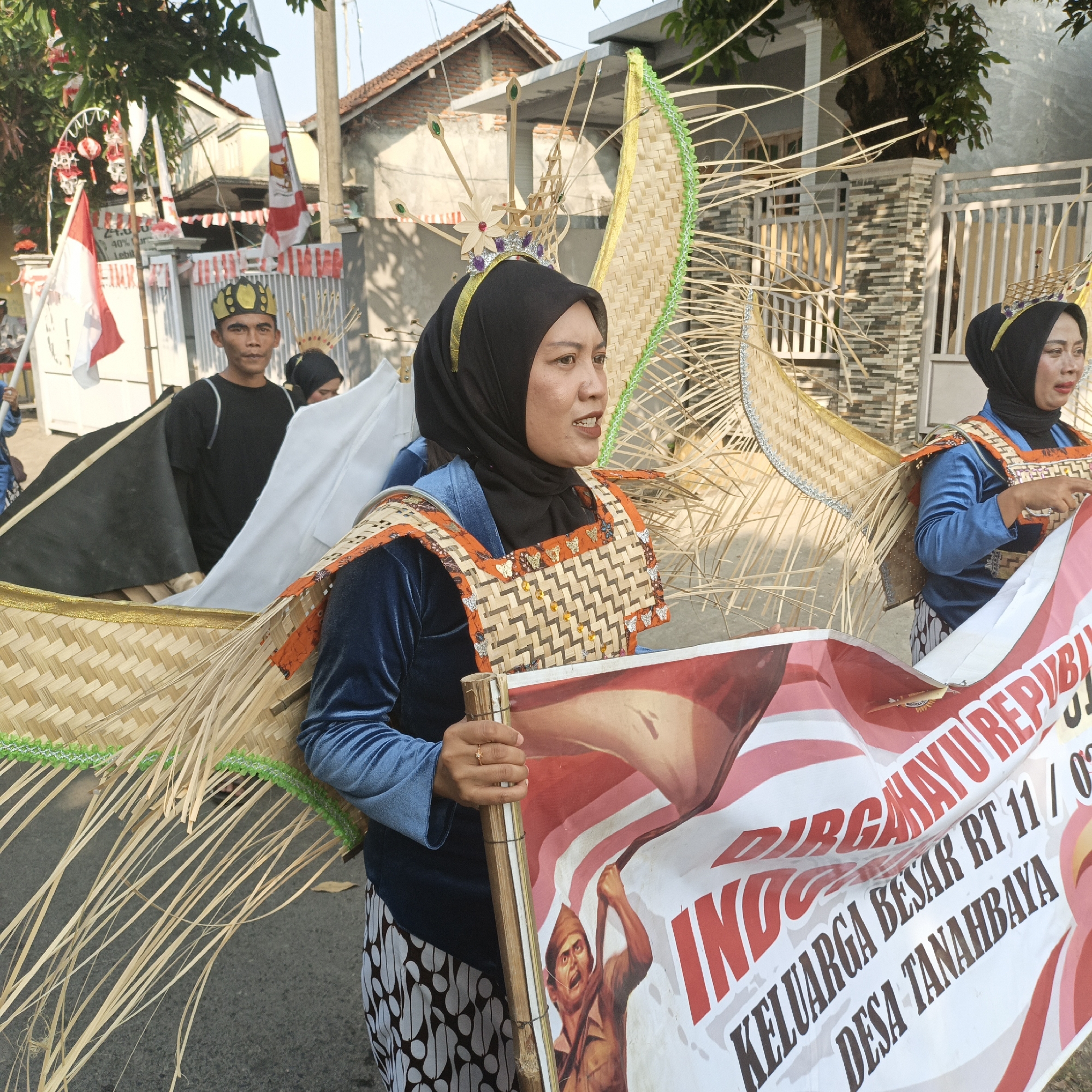 In Tanahbaya, Indonesia, a parade is underway. In the foreground, a woman wearing a blue velvet dress and a black hijab stands with a banner. The banner, held by a bamboo pole, features a patriotic design with the Indonesian flag's colors and text that includes "INDONESIA" and "KELUARGA BESAR RT 11 / 02 DESA TANAHBAYA". She wears a decorative headdress made of woven material and straw. To her left, another woman, similarly adorned with a woven headdress and headdress, is visible. Behind them, a man in a black t-shirt and a traditional cap looks forward. The background shows houses, a fence, trees, and several Indonesian flags, suggesting a celebration of national pride.