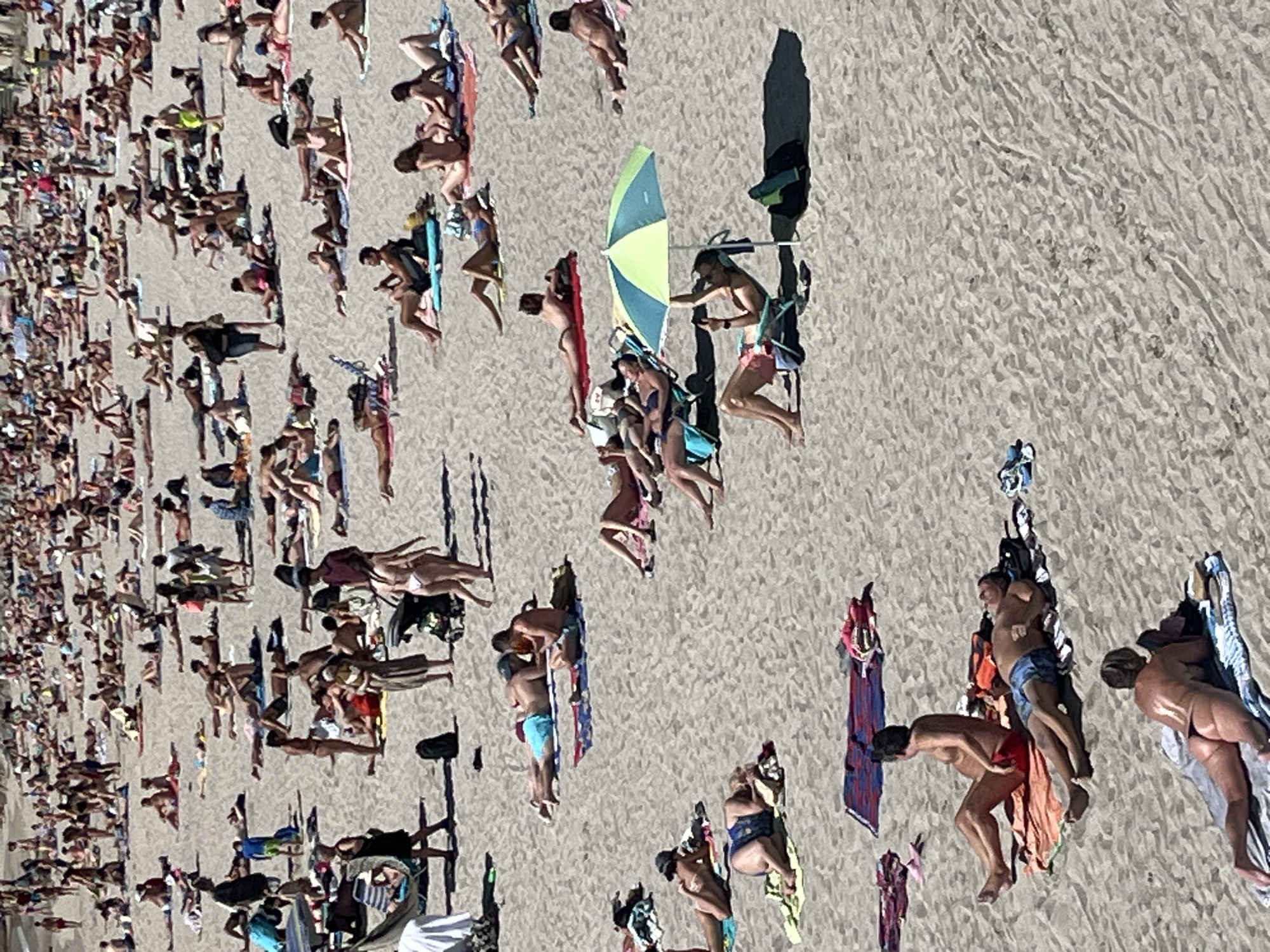 This is an aerial photograph of a crowded beach scene, likely in A Coruña, Spain, given the context. The sand is a light brown, textured with footprints and the shadows cast by the sun indicate it is midday. The sun appears to be bright, suggesting clear weather.
The primary subjects are a large number of people sunbathing and relaxing on the sand. They are spread across the frame, some lying on towels, others sitting or standing. Many are dressed in swimwear, while others are nude. There are various beach accessories visible, including towels of different colors and patterns, and a green and blue umbrella providing shade. Some individuals are interacting, with a few couples appearing to be close. The overall atmosphere seems relaxed and leisurely, typical of a busy beach on a sunny day.
Notable details include the diverse range of positions people are in, suggesting a variety of activities from sleeping to socializing. The shadows are long and sharp, reinforcing the midday timing. The high angle of the shot emphasizes the sheer number of people present, creating a mosaic of bodies and colorful towels against the sandy backdrop. No visible text is present in the image.