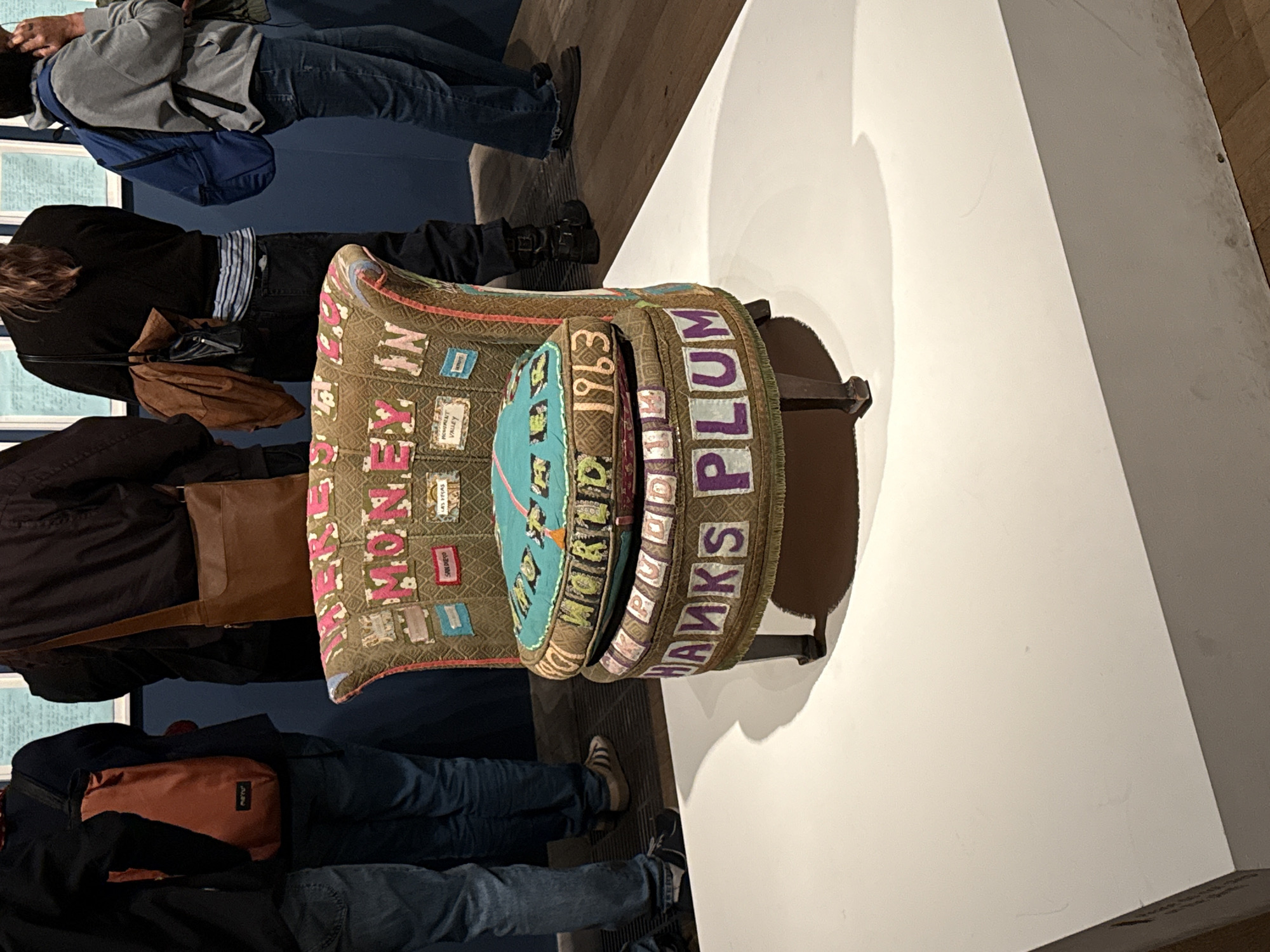 The image depicts an art installation featuring an embellished armchair on display, likely in a gallery or museum setting in London, United Kingdom, given the presence of other individuals viewing the piece.
The armchair is elaborately decorated with appliqués, embroidery, and text; the words "MONEY", "WORLD 1963", and "THANKS PLUM" are visible. Several people stand around the artwork, examining it. The environment includes neutral-colored walls and flooring. The lighting is primarily provided by overhead gallery lights.