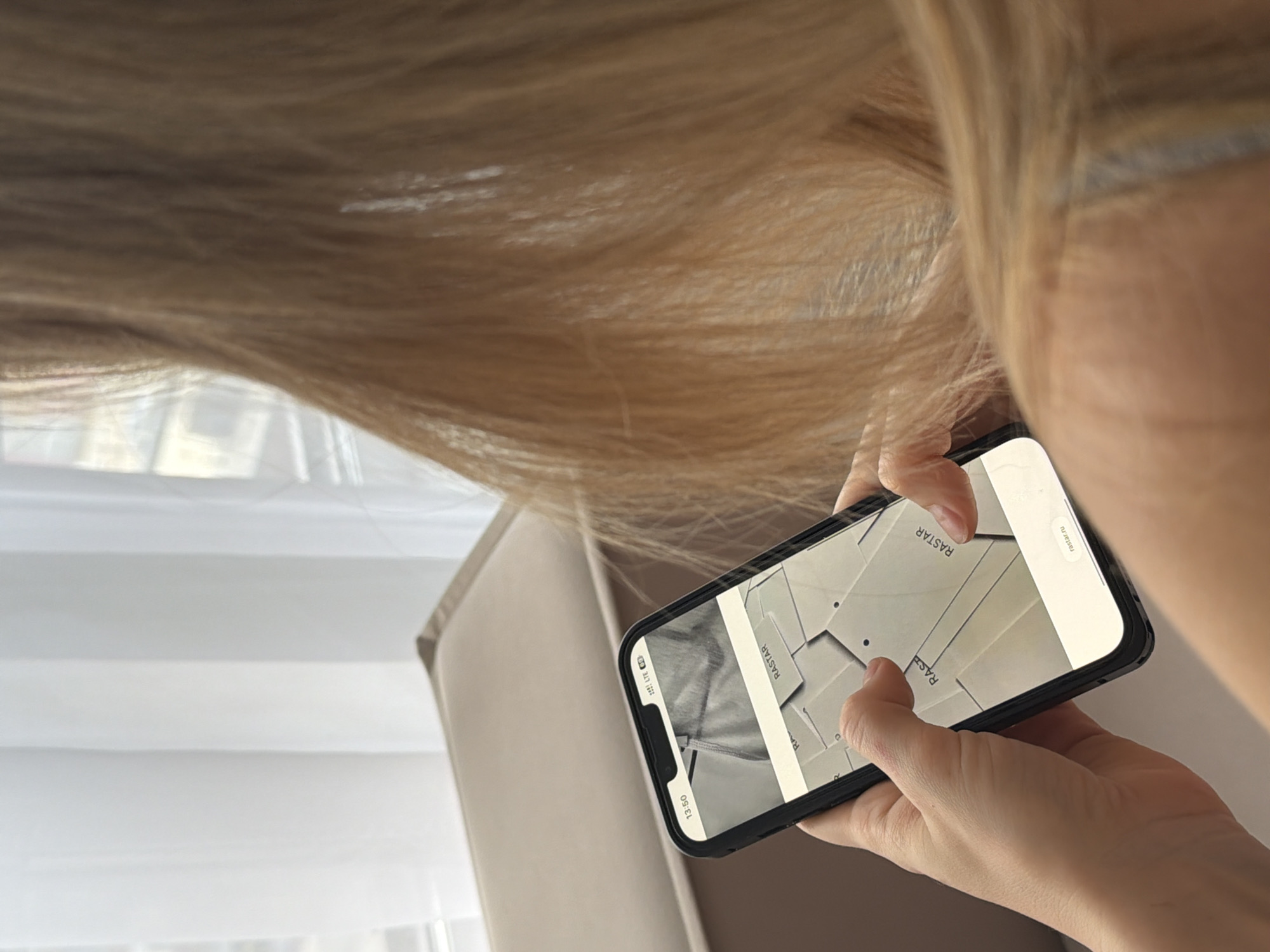 A person is holding a smartphone in their hands, displaying a pattern, possibly for sewing, with the name "RASTAR" visible on the screen. The background includes fair-colored hair, a window with blinds, and part of a neutral-colored chair or couch. No specific location in Khimki, Russia, is evident in the image.