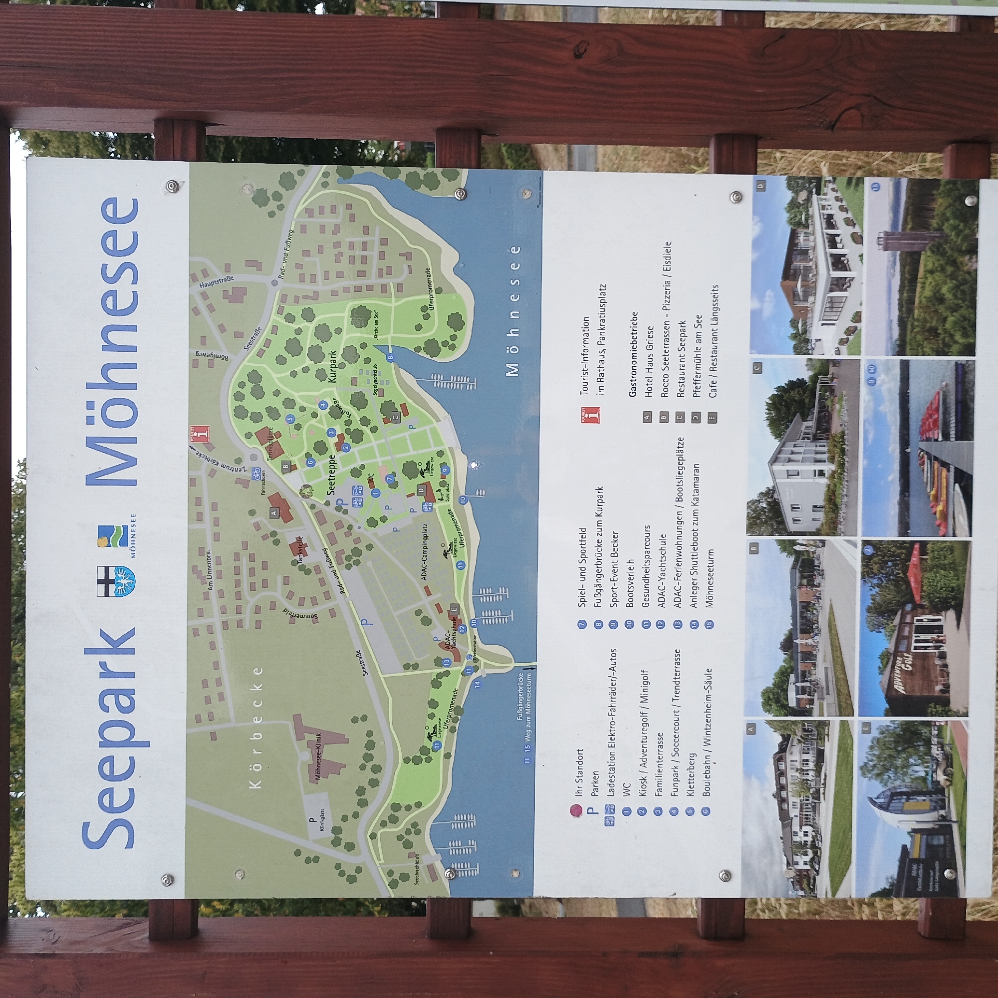 The image shows a sign in Möhnesee, Germany, with the title "Seepark Möhnesee." The sign features a map of the park, illustrating various amenities and locations such as the Möhnesee-Klinik, Kurpark, Seetreppe, and harbor. Below the map, a legend lists facilities like parking, charging stations, restrooms, adventure golf, and various restaurants. The sign also includes several photographs of buildings and attractions within the Seepark, such as hotels, restaurants, an adventure golf facility, and a view of the lake with watercraft. The sign is mounted on a wooden post and set against a natural background with trees and dry grass.