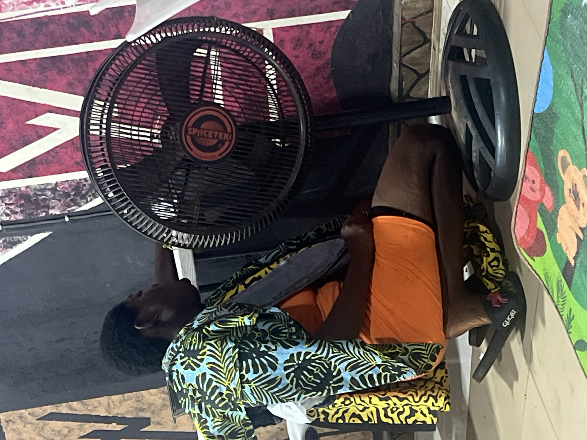 This image, taken in Obosi, Nigeria, depicts a person relaxing indoors in front of a standing fan. The person, with dark skin and short dark hair, is seated or reclining on a chair or stool, wearing a vibrant blue, black, and yellow patterned short-sleeved shirt and orange shorts or a wrapped garment. Their head is tilted back, facing the fan, indicating they are seeking relief from heat.
A black "SPACETEK" brand standing fan is positioned to the left of the person. The chair or stool beneath them is covered with a distinctive yellow and black wavy patterned fabric. On the floor nearby, a black slipper with the brand "GUCCI" visible in white lettering rests.
The background wall features varied decor. To the left, behind the fan, the wall is a dark red or maroon with sharp white geometric patterns. To the right, behind the person, the wall is a lighter tan or beige and adorned with colorful, hand-painted murals depicting cartoon-like animals, including a bear. The ambient indoor lighting suggests a relaxed setting, possibly in a home or a versatile community space.