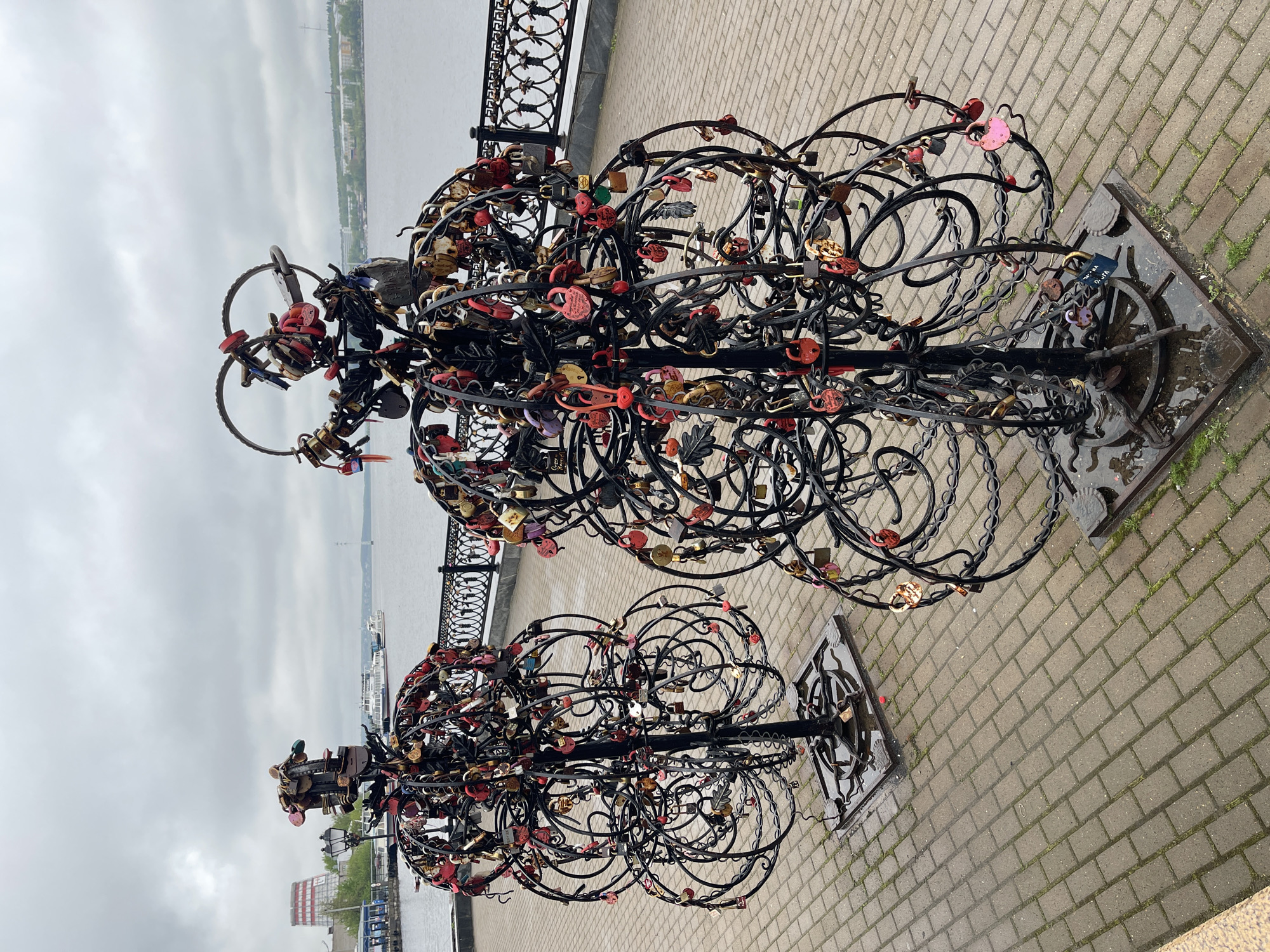The photograph depicts two metal "love trees" laden with padlocks, standing on a brick walkway in Izhevsk, Russia. The background includes a river and a wrought-iron fence. The sky is overcast. The trees are densely covered with locks of various colors and sizes, attached by couples as a symbol of their love. The bases of the trees are square and metallic. The walkway is wet in places, suggesting recent rain.