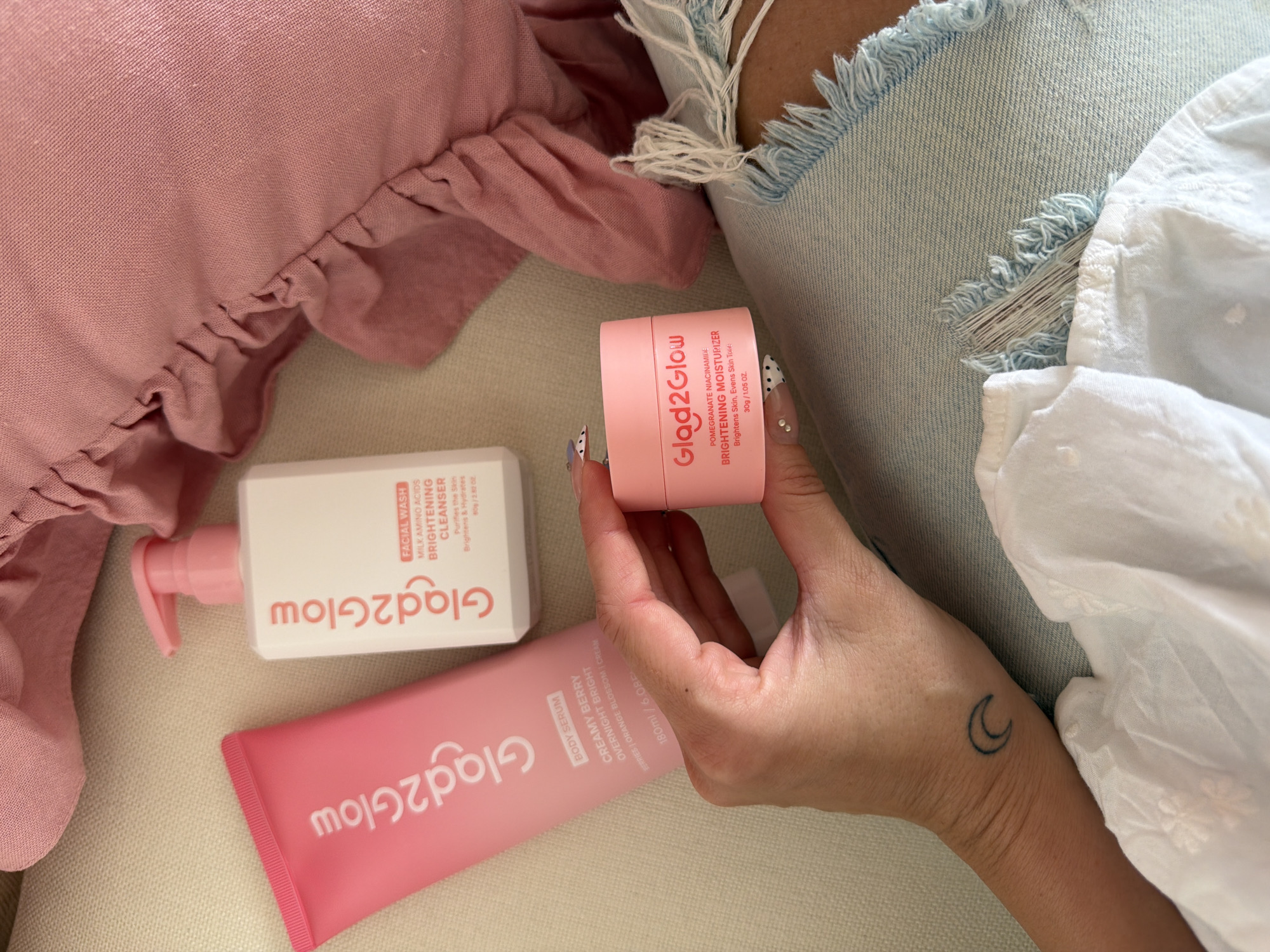 This image presents a flat-lay scene, featuring three Glag2Glow brand skincare products against a casual setting. A pink pillow with frills is visible in the upper left corner, adjacent to a pair of distressed denim shorts and a white textured shirt or cloth.
The skincare products are: a white bottle of Facial Wash Brightening Cleanser, a pink tube of Creamy Berry Body Serum, and a pink jar of Brightening Moisturizer, held in a person's hand. The products' branding is oriented to be read from top to bottom, indicating the intended viewing perspective.
A person's hand is holding the pink jar. The person has manicured nails with a polka dot pattern on some of the nails and a crescent moon tattoo on their wrist. The overall setting suggests a personal space, potentially indoors.