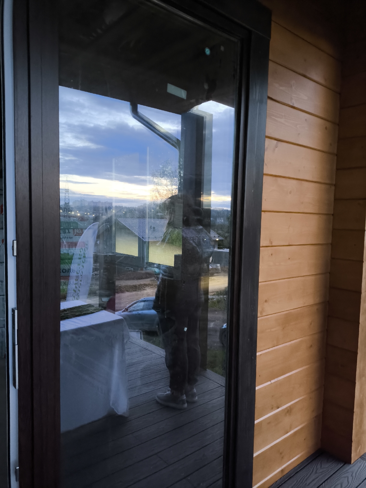 A reflection in a glass door shows a person standing on a deck with a table covered in a white cloth and a green runner. Beyond the deck are cars and a two-story building, possibly under construction, with a construction fence visible. The sky is dusky with a hint of orange on the horizon, suggesting twilight. The door frame is dark and wooden, and the wall adjacent to the door is made of light-colored horizontal wooden planks. The person reflected in the glass appears to be wearing dark clothing and light-colored shoes.