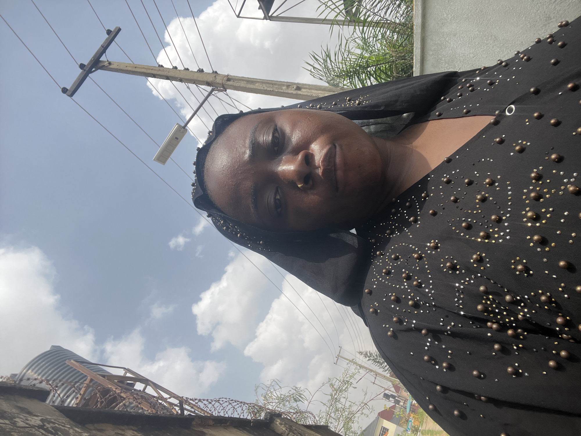 A woman wearing a black beaded garment is centered in the image. The background features a cloudy blue sky, a utility pole with wires, and a palm tree. A corrugated metal structure with barbed wire is visible on the left side. The location is in Abuja, Nigeria.