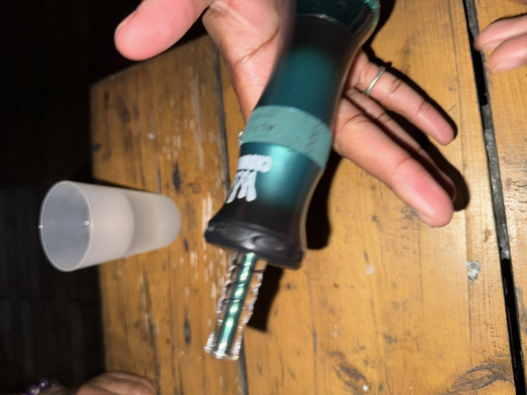 An individual holds a dark green vaporizer with a clear stem over a wooden table. A translucent cup sits next to the vaporizer on the table. The individual's fingers are visible, with a ring on one finger. The table appears to be indoors, with a dark background. The location is unknown.