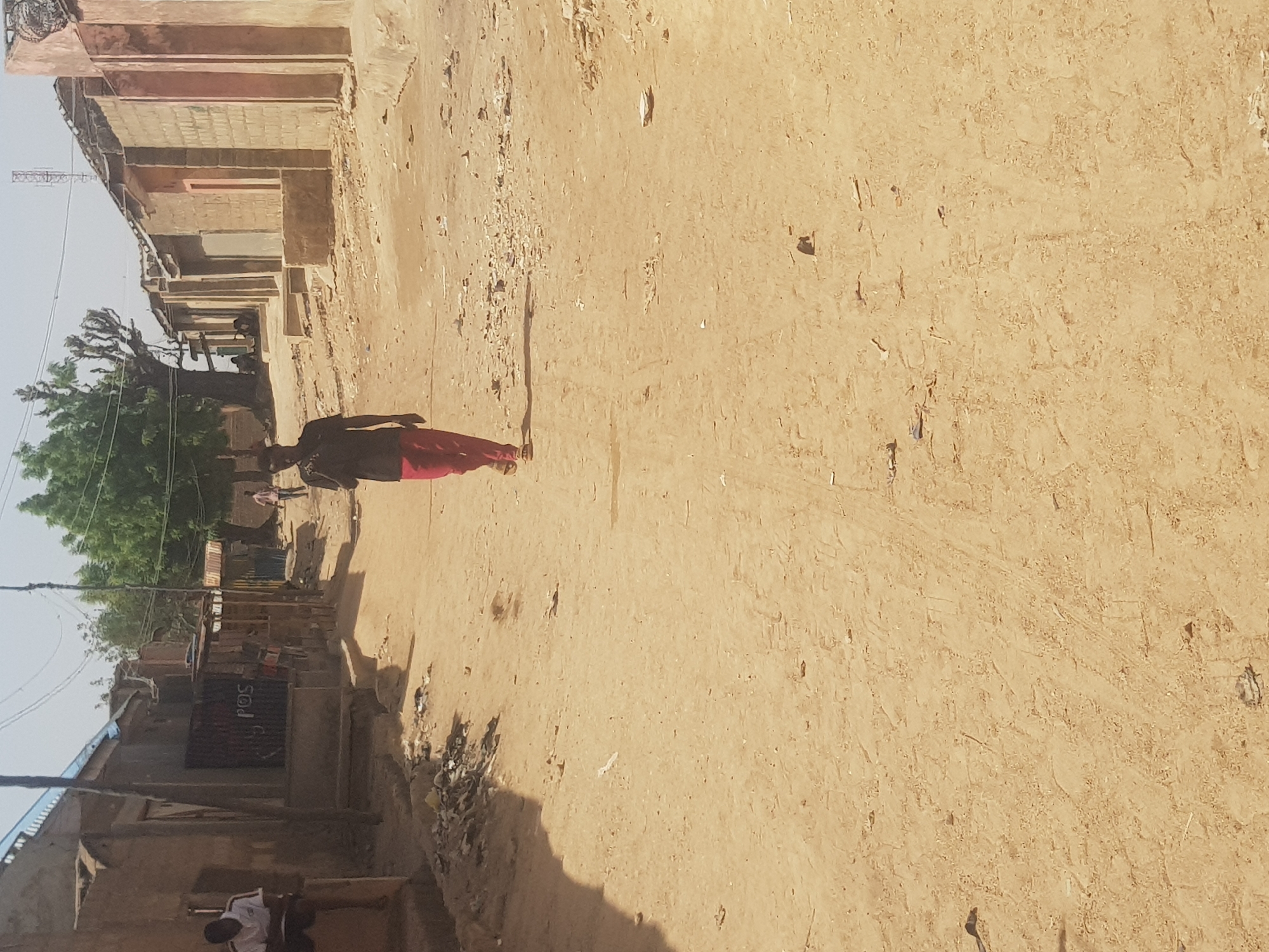 A dusty, sun-baked street scene in Dabawa, Nigeria. A person wearing dark clothes and red trousers is walking along the sandy road. Buildings of simple construction line the sides of the road, with a POS sign visible on one of them. A tree with green foliage is located near the left side of the frame. Other people can be seen in the background. The overall impression is one of a warm, arid environment.