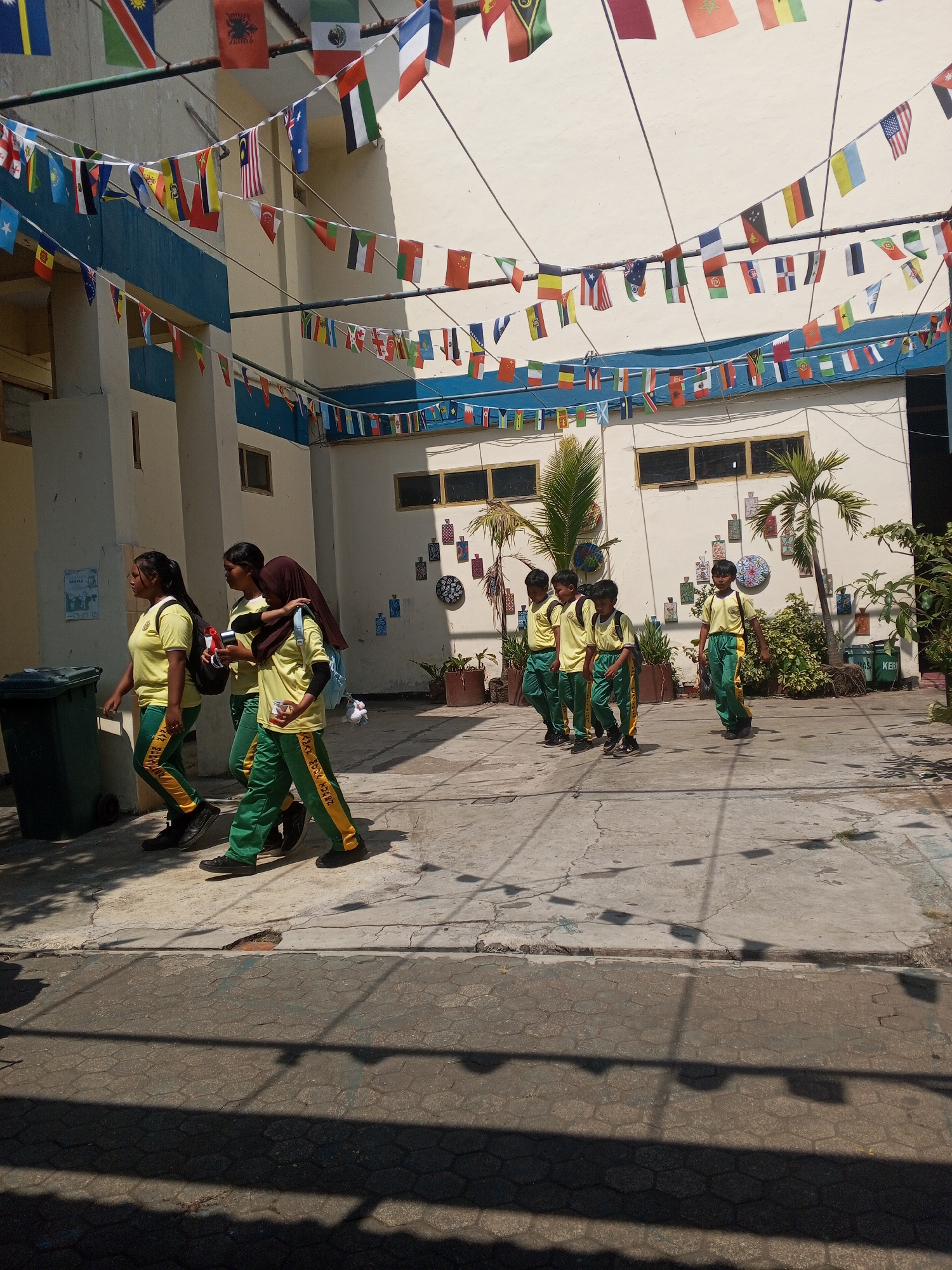 In Turen, Indonesia, a group of students, predominantly in school uniforms, walk along a paved courtyard. The students are wearing yellow shirts and green pants with yellow stripes. Some of the students are carrying backpacks, and one student in the foreground on the right is carrying a red and white cup. The environment is outdoors, with a building in the background adorned with numerous international flags strung across wires. The building has a light-colored facade with blue trim and windows. In the courtyard, there are potted plants and a dark green trash bin. The scene is illuminated by sunlight, casting shadows on the ground.