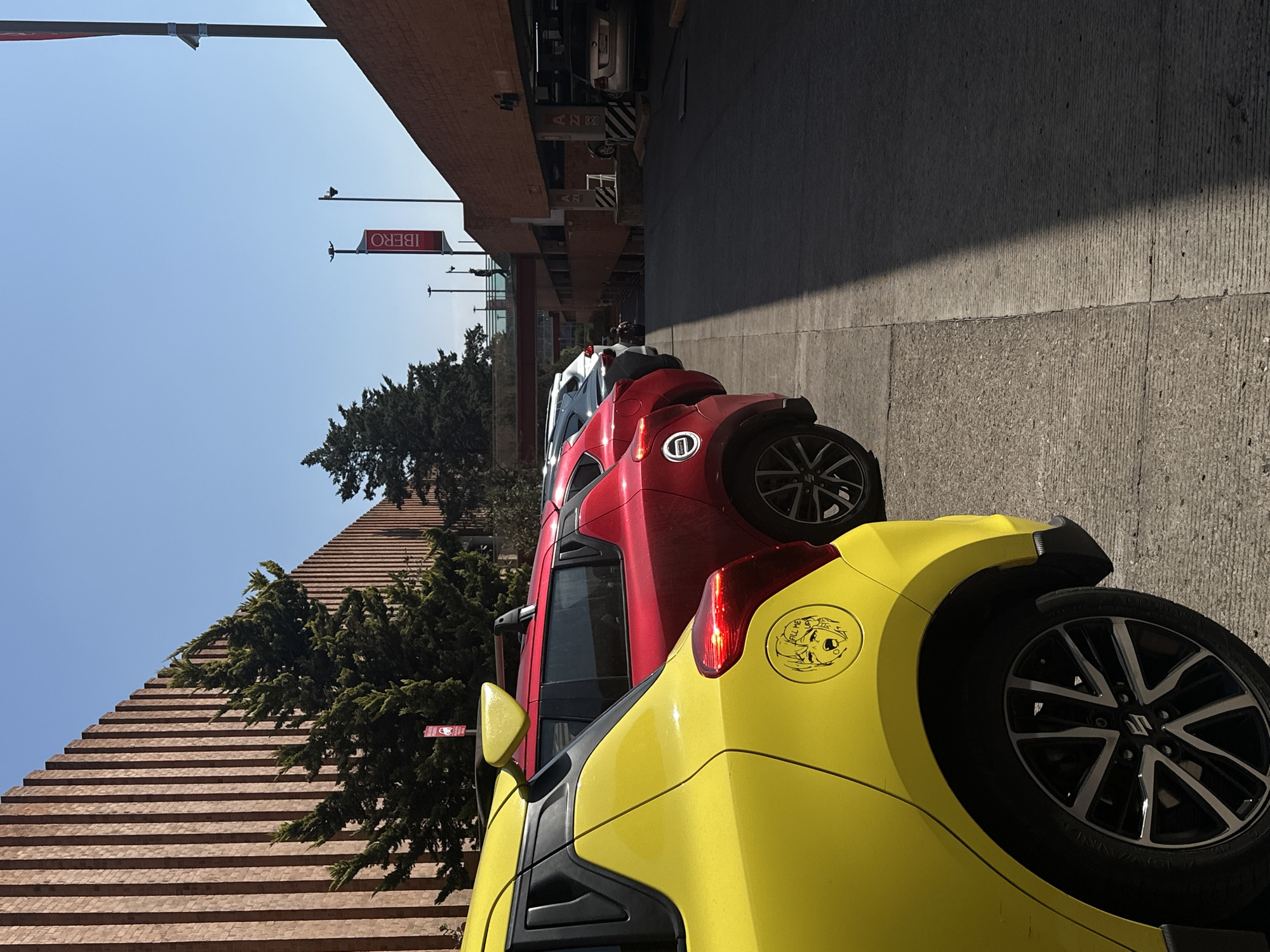 The image shows a row of parked cars on a street in Mexico City, Mexico. The cars are parked on a concrete surface, likely a sidewalk or parking lane, next to a building with a distinctive stepped, brick-like facade. A sign with the word "IBERO" is visible on a lamppost. The closest car is yellow and has a sticker on its rear fender. Behind it is a red car, followed by a white car. The sky is clear and blue. The photo appears to be taken from ground level, focusing on the parked cars and the surrounding buildings and street.