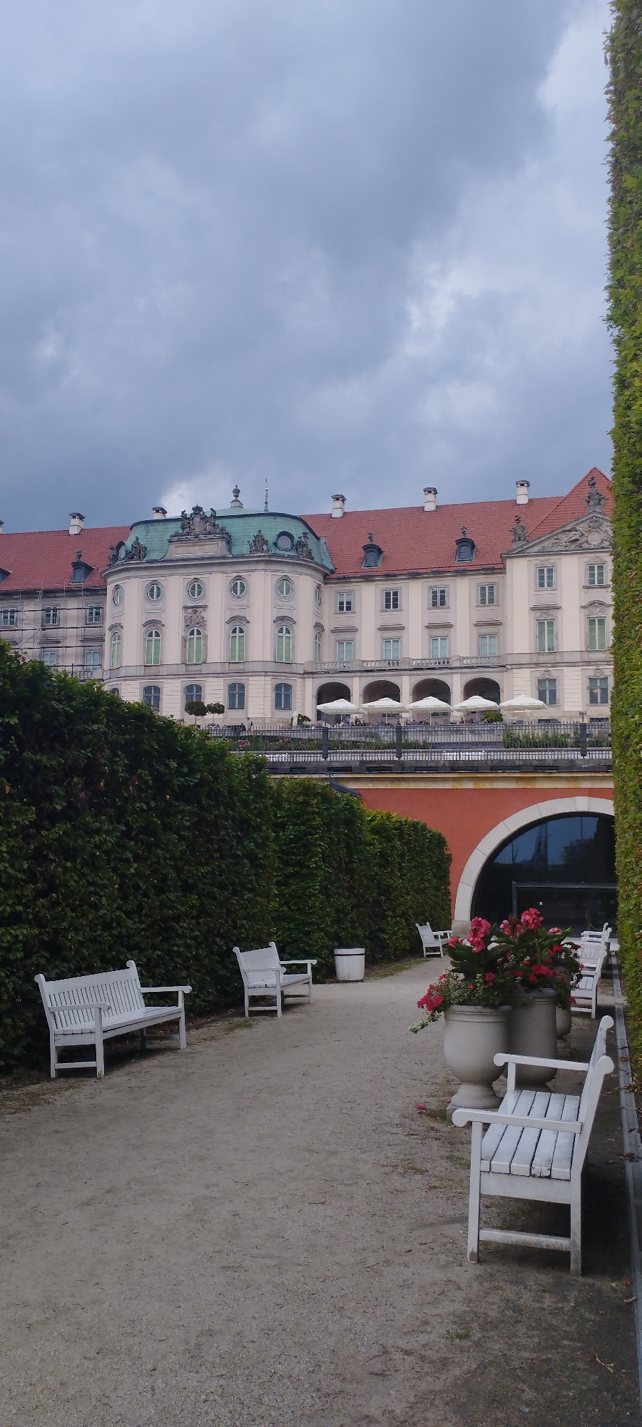 The image depicts a section of the Royal Castle in Warsaw, Poland, on a cloudy day. The castle, with its pale facade and red tiled roof, is visible in the background. In the foreground, a manicured garden with a gravel path and white benches is present. A tall, dense hedge lines the left side of the path, and a large ivy-covered wall is on the right. Two white wooden benches are positioned along the path, facing the castle. In the middle ground, planters with pink flowers and a large arched entrance are visible. The sky is overcast and grey. No people are clearly discernible in the image.