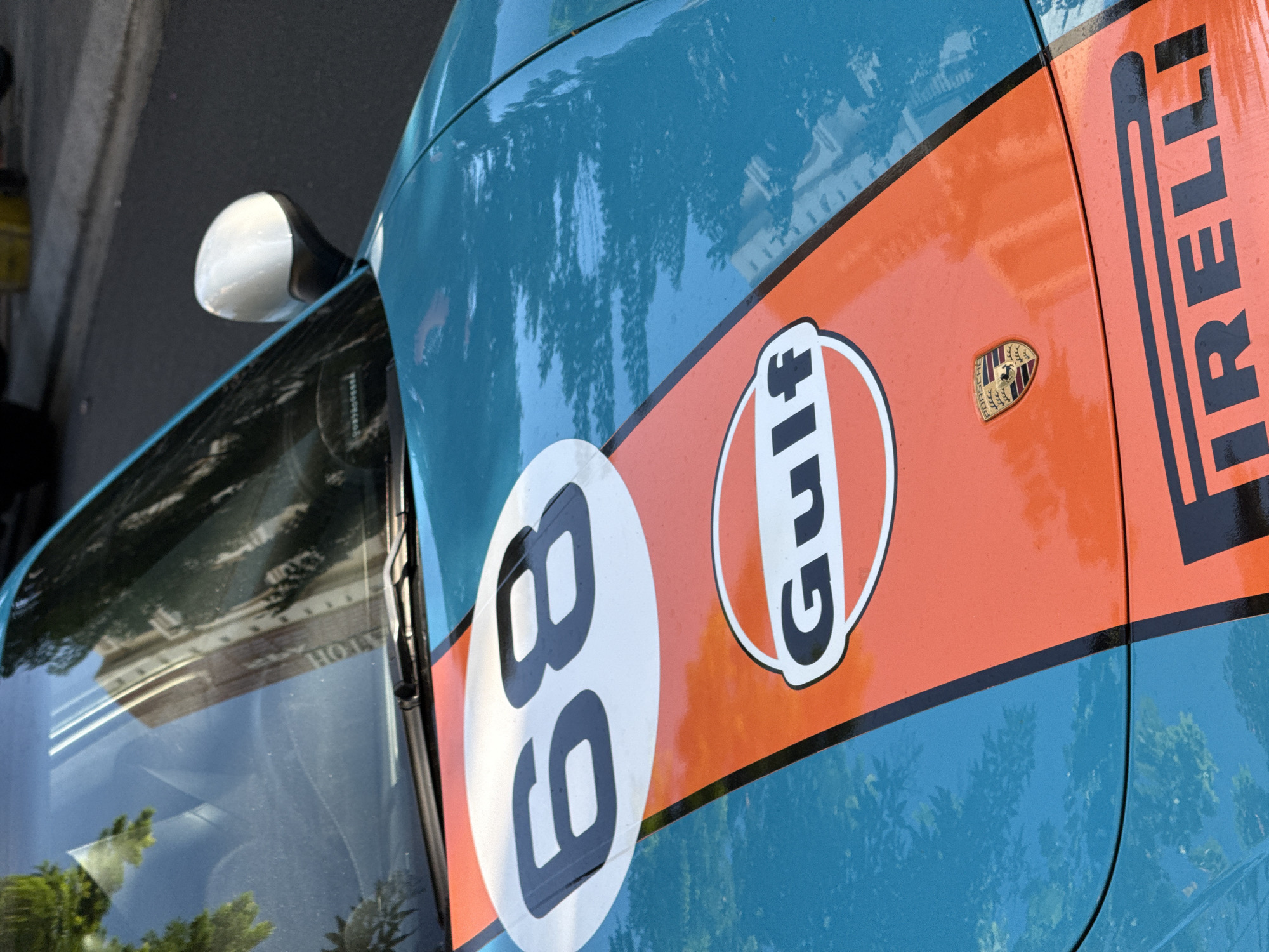 The image shows a close-up of a car with Gulf racing livery, featuring an orange stripe with the number 68 in a white circle, a Gulf logo, a Porsche emblem, and a Pirelli logo. The car is turquoise blue. Reflections of trees and a building are visible on the car's surface. The location cannot be determined solely from the image.