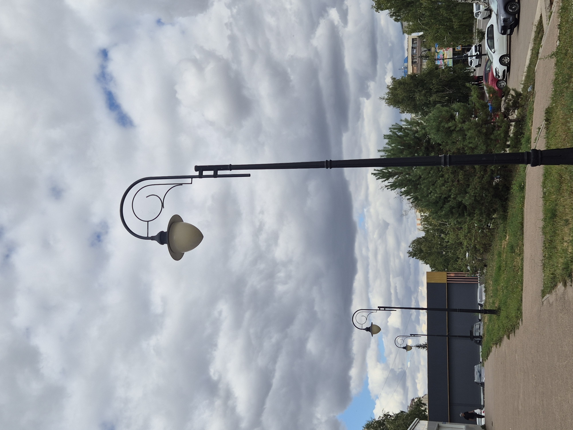 This is a medium-shot, eye-level, vertical photograph taken on a cloudy day, likely in Kokshetau, Kazakhstan. The foreground features a paved walkway on the left and a grassy area on the right. A black lamppost with an ornate curved arm and a pale, possibly glass, lampshade stands prominently in the center of the frame. Further down the walkway, two more similar lampposts are visible, receding into the distance. Beyond the walkway and lampposts, a modern building with a dark gray facade and a yellow-trimmed section is visible. Several parked cars, including a white SUV and a dark gray SUV, are present. Trees, including evergreen and deciduous varieties, line the background, with more buildings and a portion of what appears to be a billboard visible further beyond. A single person, dressed in dark clothing, walks away from the camera on the far left of the walkway. The sky is filled with heavy, gray, cumulus clouds, with hints of blue sky visible in the upper left corner.