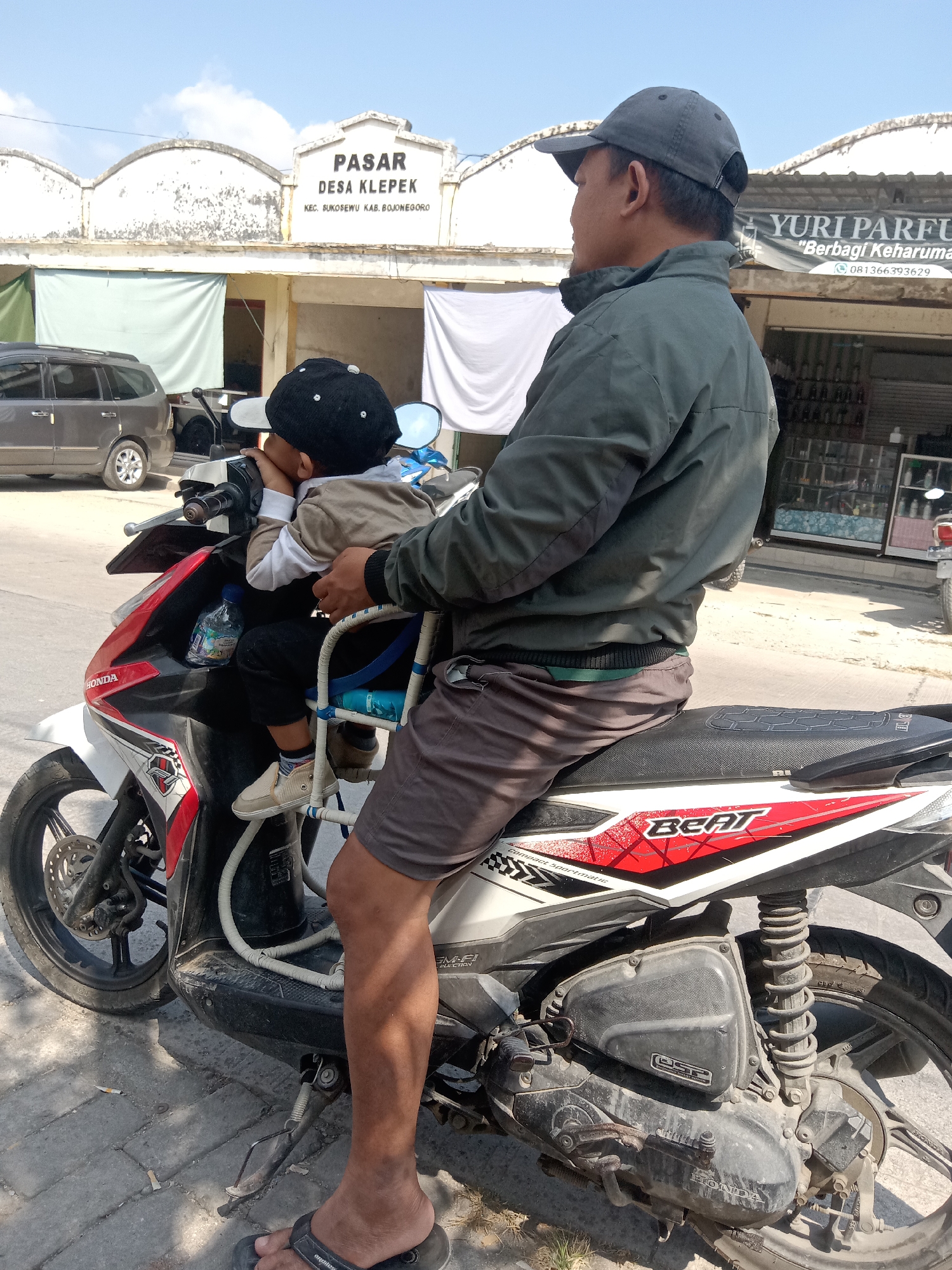 The image depicts a man and a young child on a Honda Beat motorcycle parked on a paved street in front of buildings that appear to be market stalls or shops. The man, wearing a gray baseball cap and a green jacket, is seated on the motorcycle, with the child in a blue and white child seat attached to the front of the motorcycle. The child, wearing a black baseball cap and a tan and white shirt, is leaning on the handlebar. The buildings in the background have signs with text, including "PASAR DESA KLEPEK KEC. BUKOSEWU KAB. BOJONEGORO" which indicates the location is the Klepek village market in Bojonegoro Regency, Indonesia. One of the shops has a sign that reads "YURI PARFUM". A dark gray SUV is parked to the left of the motorcycle. The lighting suggests it is daytime with a clear sky.