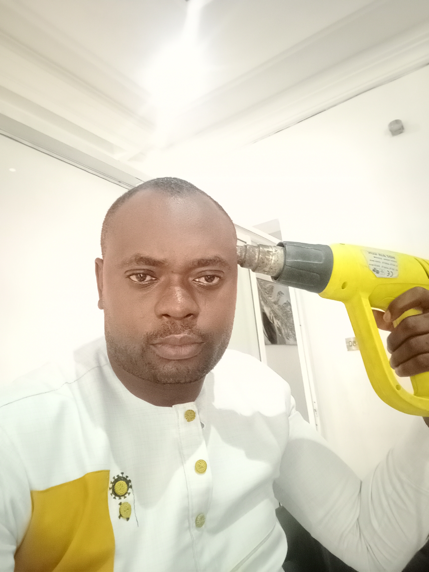 A man is holding a yellow heat gun pointed at his head. He has dark skin, a shaved head, and is wearing a white shirt with gold buttons and a gold patch. The background includes a white wall and door. There is no evidence in the image to indicate that this image was taken in Gwarinpa, Nigeria.