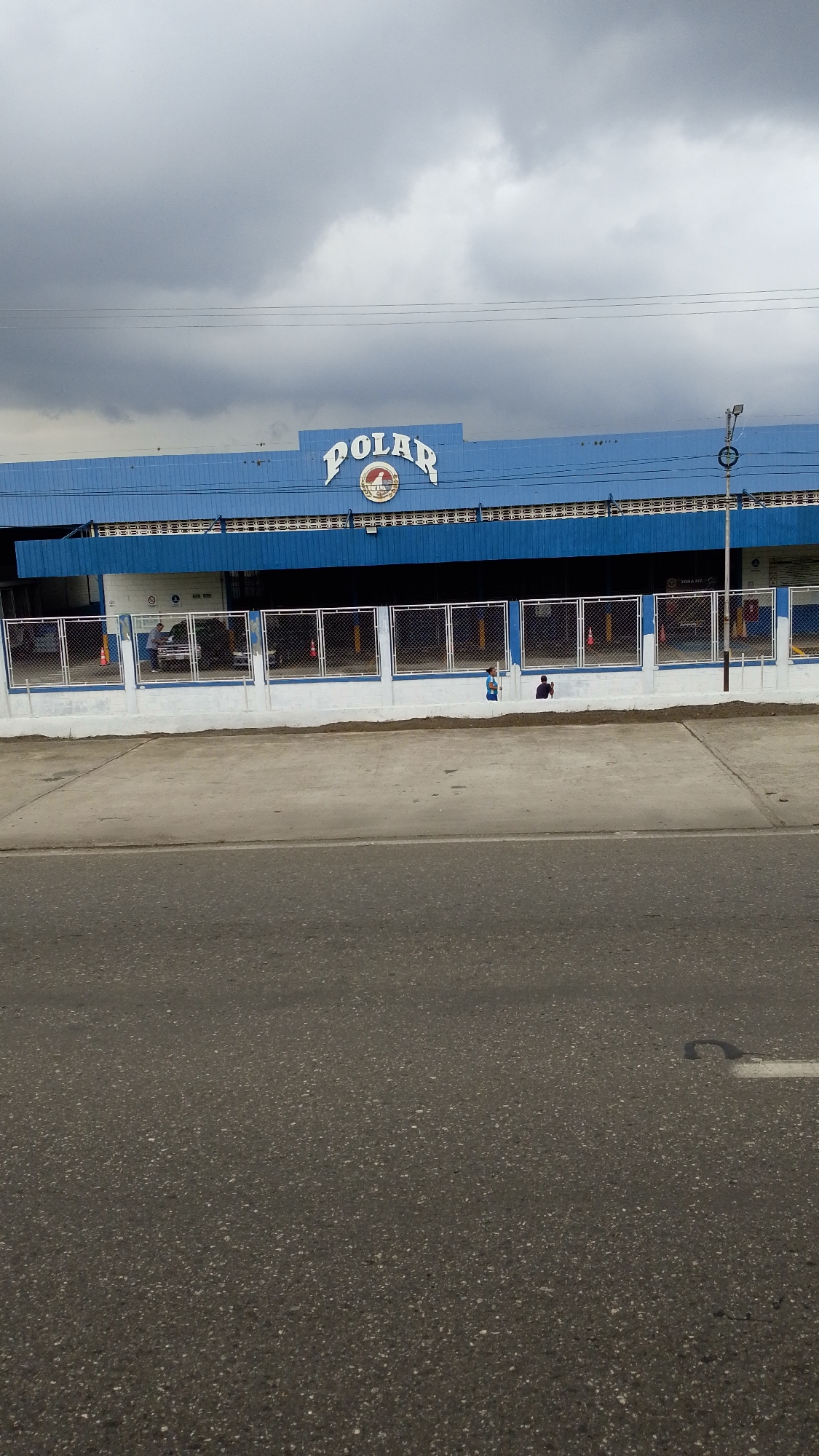 The image shows the exterior of the Polar Brewery in Tinaquillo, Venezuela. The building is a light blue, and the word "POLAR" is prominently displayed on the top. The front of the building has a white concrete wall, topped with a chain-link fence. Behind the fence, there are vehicles and what appear to be orange traffic cones. On the sidewalk alongside the fence are two indistinct figures. The foreground is occupied by a grey asphalt road, with a small, curved marking visible on the surface. The sky above is overcast with grey clouds.