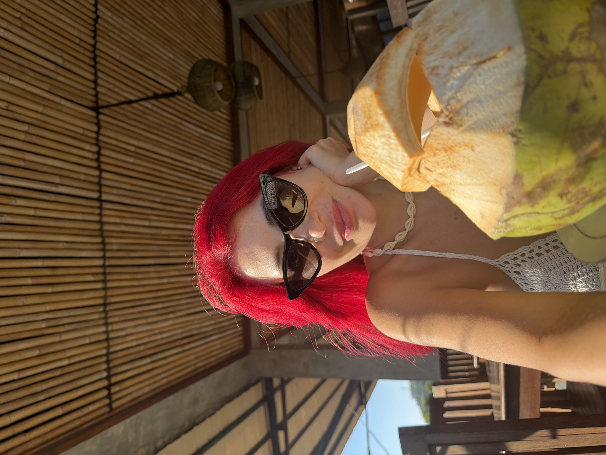 A woman with vibrant red hair and sunglasses leans her head on her hand, a tropical drink in a coconut in front of her. She is wearing a white crocheted top and a shell necklace. The background consists of bamboo walls and a thatched roof, suggesting an outdoor setting in a warm climate. The sunlight indicates it is daytime. The woman appears relaxed and happy, with a slight smile on her face. The scene evokes a vacation atmosphere, likely in a beachside resort or cafe. The location is identified as Kute, Indonesia. No visible text is present in the image.
