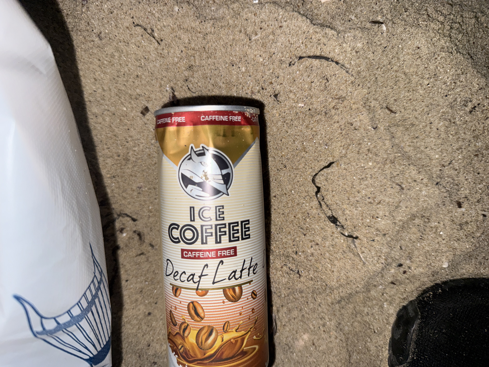 An aluminum "ICE COFFEE Decaf Latte" can lies horizontally on coarse sand in Varna, Bulgaria. The can features a gold and white design with "ICE COFFEE" prominently displayed in black text, accompanied by "CAFFEINE FREE" in a red banner below. "Decaf Latte" is written in a script font to the left. A graphic of coffee beans and a liquid splash is on the left side of the can's label, while a stylized animal head logo, resembling a cat or lion, is present on the right side of the main label. The can appears intact, with minor sandy residue on its surface.
The sandy ground is light brown/beige, composed of fine grains mixed with scattered darker specks and a few small white shell fragments. Several thin, dark, organic elements, consistent with dried seaweed or small plant debris, are visible, particularly in the lower right and central areas of the sand.
In the upper left quadrant of the image, a portion of a white, textured object, possibly a plastic bag or fabric, is visible. It features a partial blue graphic resembling a fish's tail or fin. A distinct shadow from this white object falls across the sand in the upper middle area. A dark, textured object, likely the toe of a shoe or sandal, is partially visible in the lower left corner, resting on the sand. No individuals or active interactions are depicted in the frame.