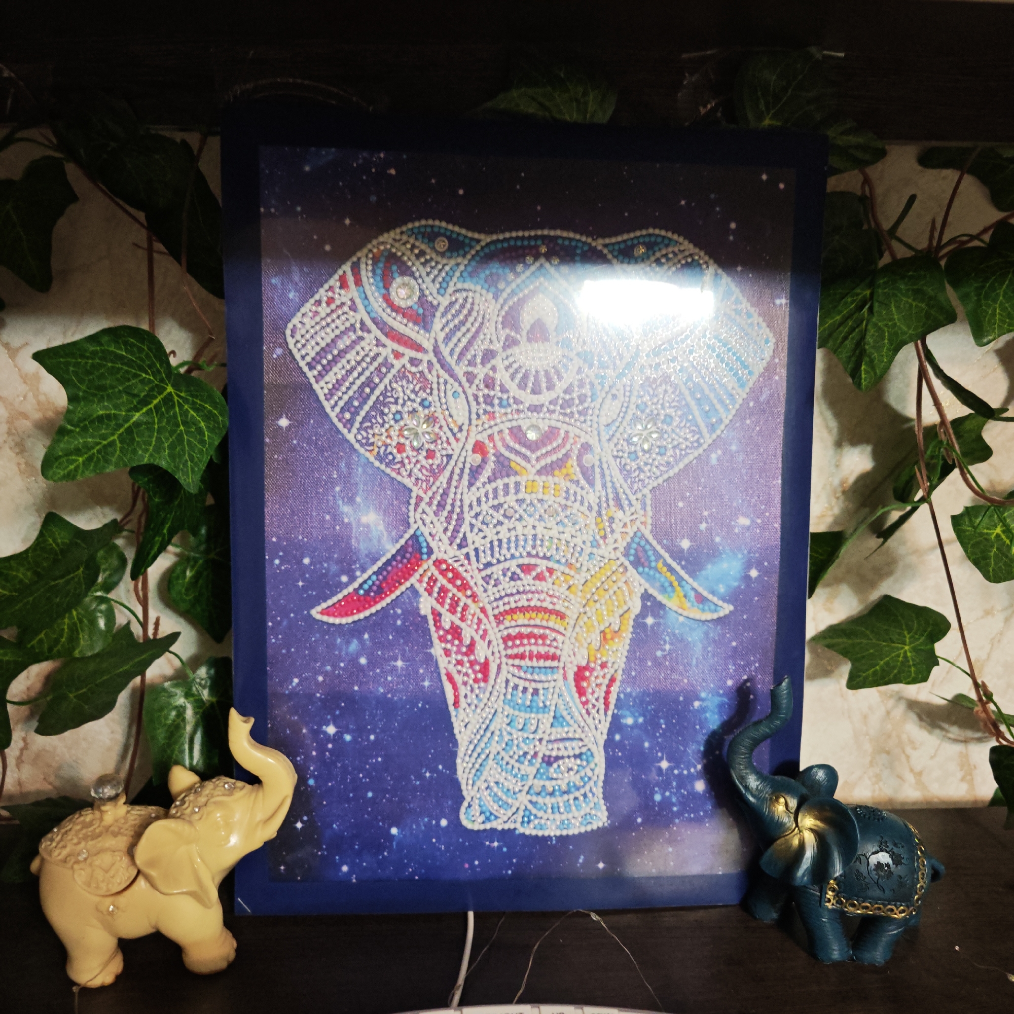 The image shows an art display featuring an elephant motif, potentially located indoors. A diamond painting depicting an elephant in colorful patterns against a blue, starry background is the central element. Flanking the painting are two small elephant figurines: one is a light yellow color with decorative elements, and the other is a darker, teal color with gold accents. Green ivy vines frame the scene on either side, adding a touch of nature to the display. The objects are situated on a dark, possibly wooden, surface that appears to be a shelf or tabletop. Based on the prompt's request, this would likely be an indoor display somewhere in the city of Rostov, Russia, but specific location information would not be available without other data.