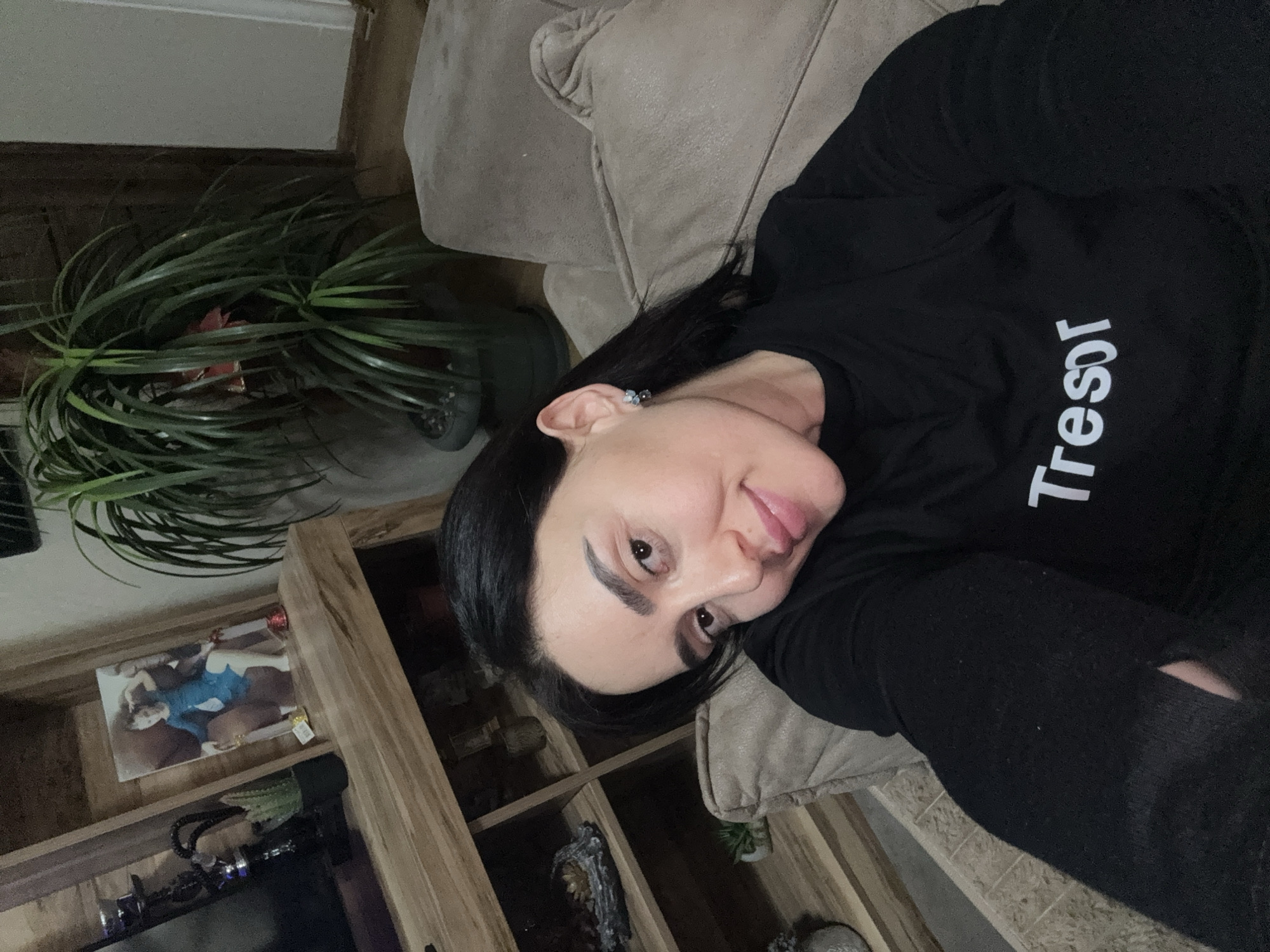 A woman with dark hair is reclining on a beige couch in what appears to be a residential setting. She is wearing a black long-sleeved shirt with the word "Tresor" printed on it in white. To her left is a wooden shelving unit filled with various items, including a photograph and decorative objects. A potted plant with long, drooping green leaves is visible behind the shelving unit. While it is impossible to confirm the location in the city of Tbilisi, Georgia based on the image alone, the scene suggests a domestic interior.