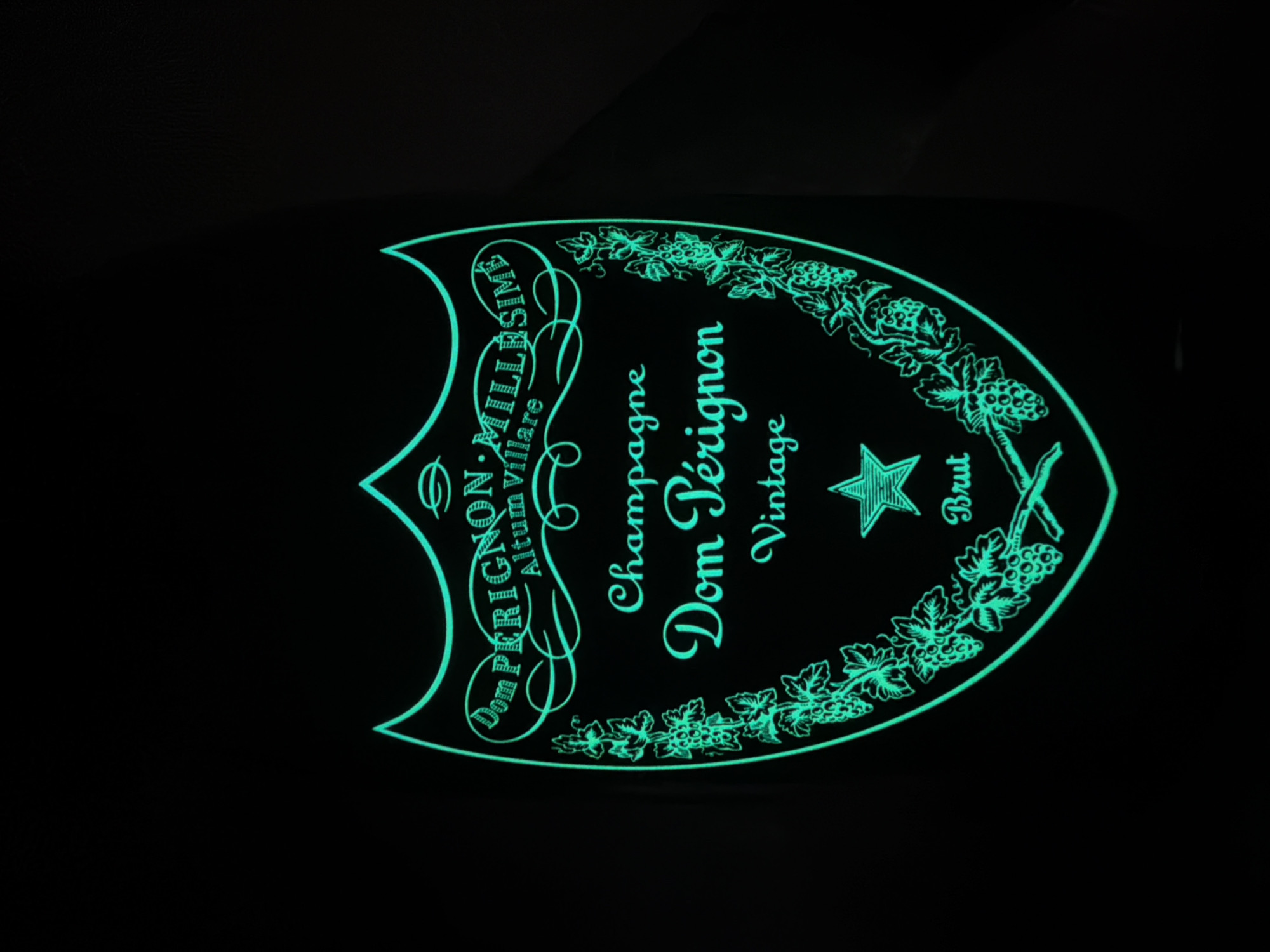 A neon green outline of a Dom Pérignon champagne label glows against a black background. The label is shield-shaped, with intricate decorative elements including grapevines and a star. Text on the label reads "Dom Pérignon Millésime," "Altum Villare," "Champagne," "Dom Pérignon," "Vintage," and "Brut." The image does not provide any information about the location in Dolno Ablanovo, Bulgaria.