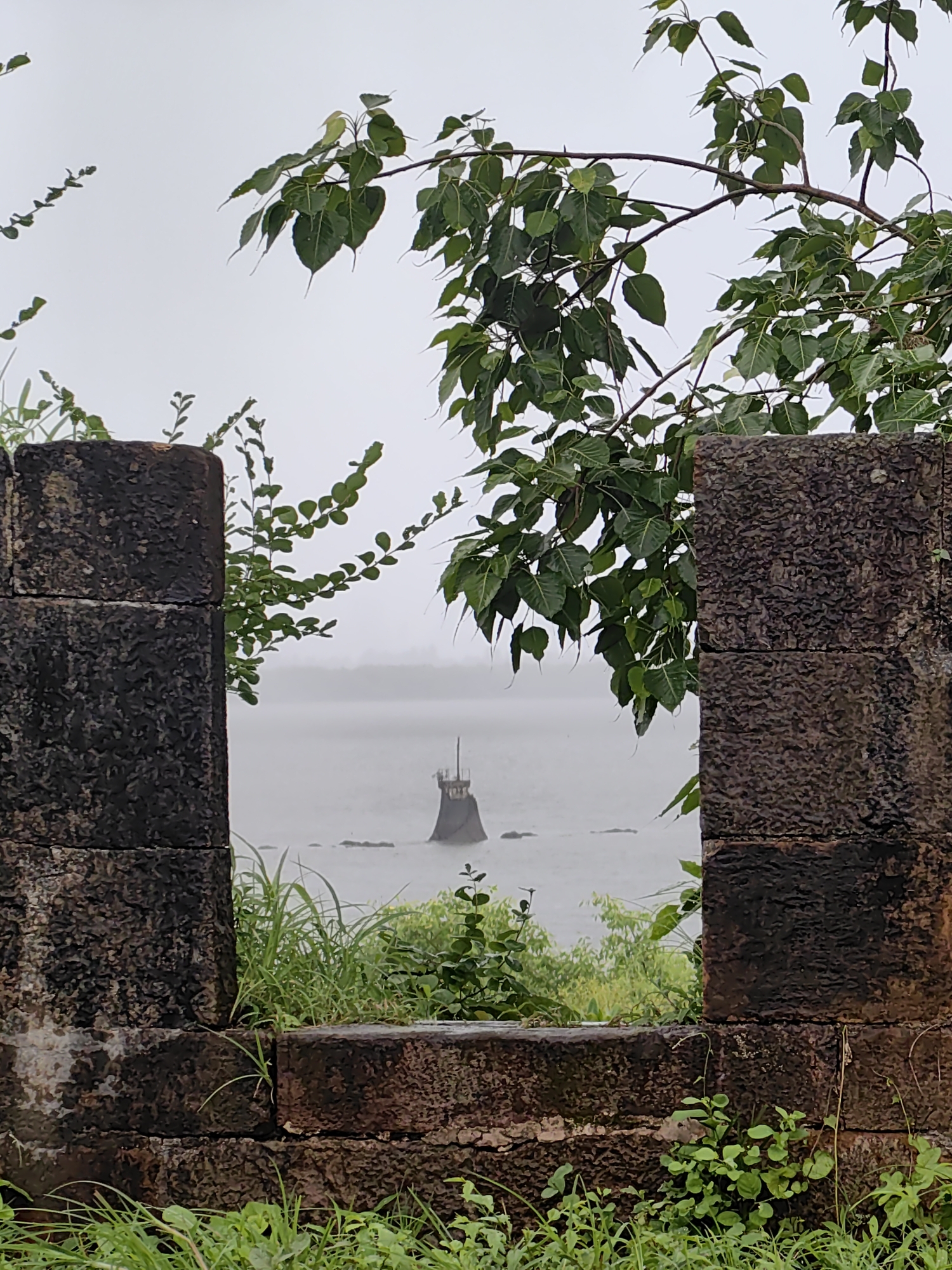 The image depicts a view through a weathered stone structure, likely part of a fort or historical ruin, towards a body of water under an overcast sky. In the foreground, two large, rough-hewn stone pillars frame the scene. Lush green grass and small plants grow around the base of the stone structure.
Through the gap between the pillars, a calm body of water, possibly the sea or a large river, is visible. In the distance, a solitary, dark, conical structure rises from the water. This structure appears to be a beacon, lighthouse, or a submerged monument. The horizon is hazy, suggesting a misty or foggy atmosphere.
The overall impression is one of age and natural overgrowth, with the ancient stonework contrasted against the vibrant greenery and the expansive, muted waterscape. The presence of the stone structure strongly suggests a historical site, and given the context of Vasai-Virar, India, it is highly probable that this scene is located at Vasai Fort, a prominent historical landmark in the region known for its ruins and proximity to the coast.