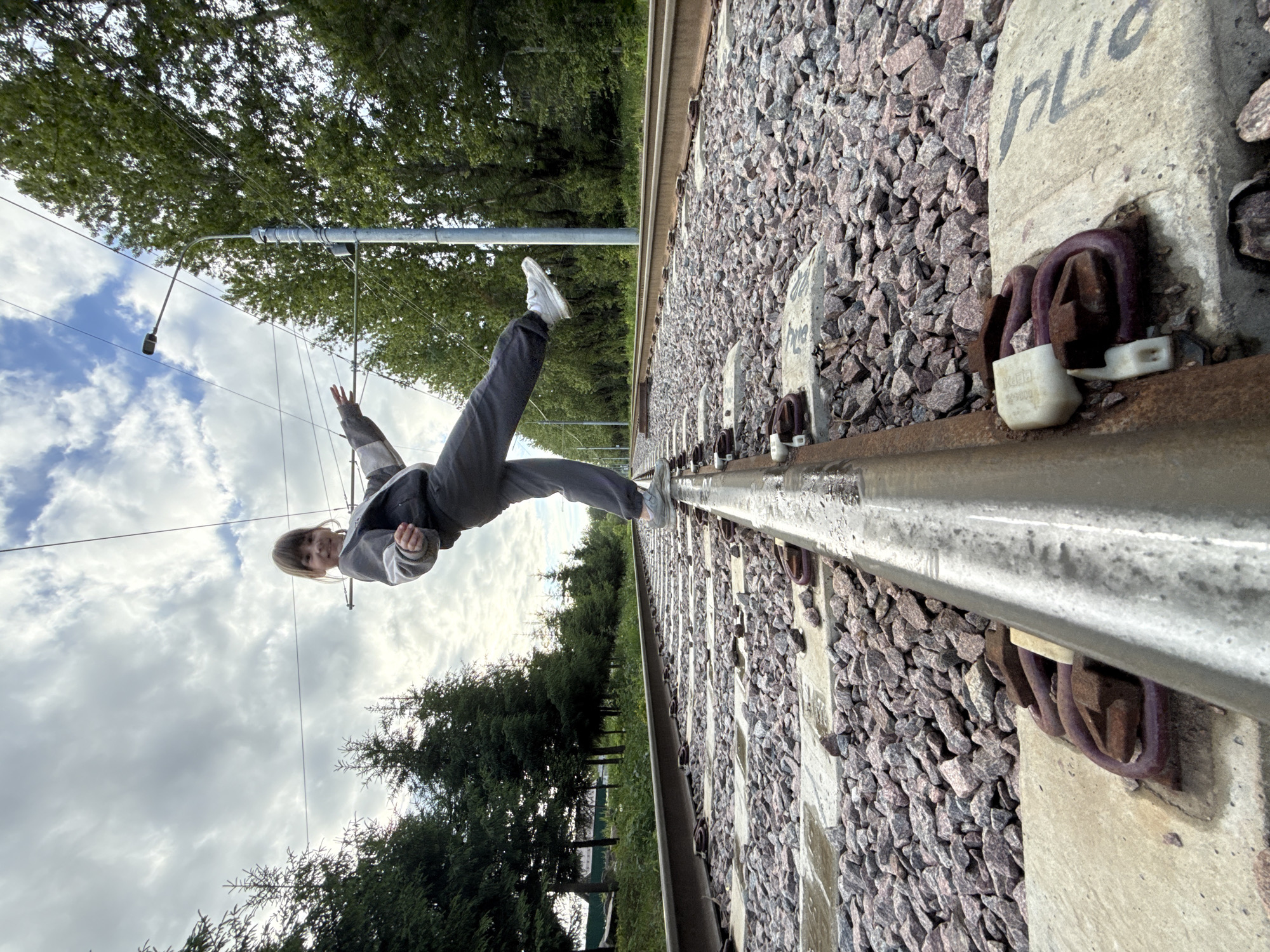 A person is balancing horizontally on a train track. The person is wearing a gray tracksuit and white sneakers. The setting appears to be outdoors, alongside a railroad track. The track runs through a green, tree-lined area. The railway ballast consists of small, angular, reddish-gray rocks. The concrete rail sleepers are visible, secured to the track with metal fasteners. In the background, overhead wires extend, likely for electric train operation. The sky is cloudy. The location may be Rusanovo, Russia, but there is no detail that proves this.