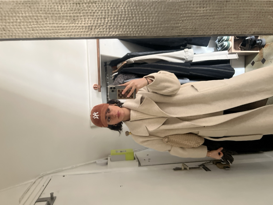 A young adult female is depicted in a mirror selfie, standing indoors within what appears to be a residential entryway or dressing area in Paris, France. She wears a brown baseball cap with a white "NY" logo, a cream chunky knit sweater, and a long, light beige wool coat. Her short, dark hair is visible below the cap, and she wears small hoop earrings. She holds a smartphone with a patterned case in her right hand, positioning it for the photograph, while her left arm supports a woven light-colored bag and grasps a pair of dark gloves. The background, reflected in the mirror, shows plain white walls, a white door to the far left, and a light green and white utility box or electrical panel. To the right, a clothes rail displays various garments, including coats and striped shirts. Below the rail, a small wooden shelf holds a pair of dark green and white sneakers and a pair of shiny black dress shoes. The floor is tiled with a geometric pattern in light and dark brown tones, consistent with traditional Parisian apartment flooring. The mirror's frame on the right side exhibits a textured, possibly hammered metal, finish.