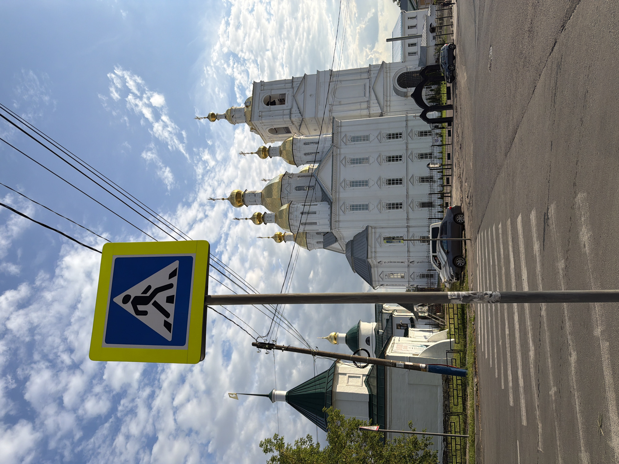 The image shows a pedestrian crossing sign in the foreground, with a large white church with golden domes in the background. The church appears to be a cathedral or a significant religious building. There are cars parked on the street near the church, and some power lines are visible overhead. The sky is blue with scattered clouds, suggesting it is daytime. The street is paved, and the pedestrian crossing is clearly marked with white stripes. The overall scene suggests a public space in a city, likely with historical or religious significance. The architecture of the church indicates a Russian Orthodox style. The location is identified as Arzamas, Russia.