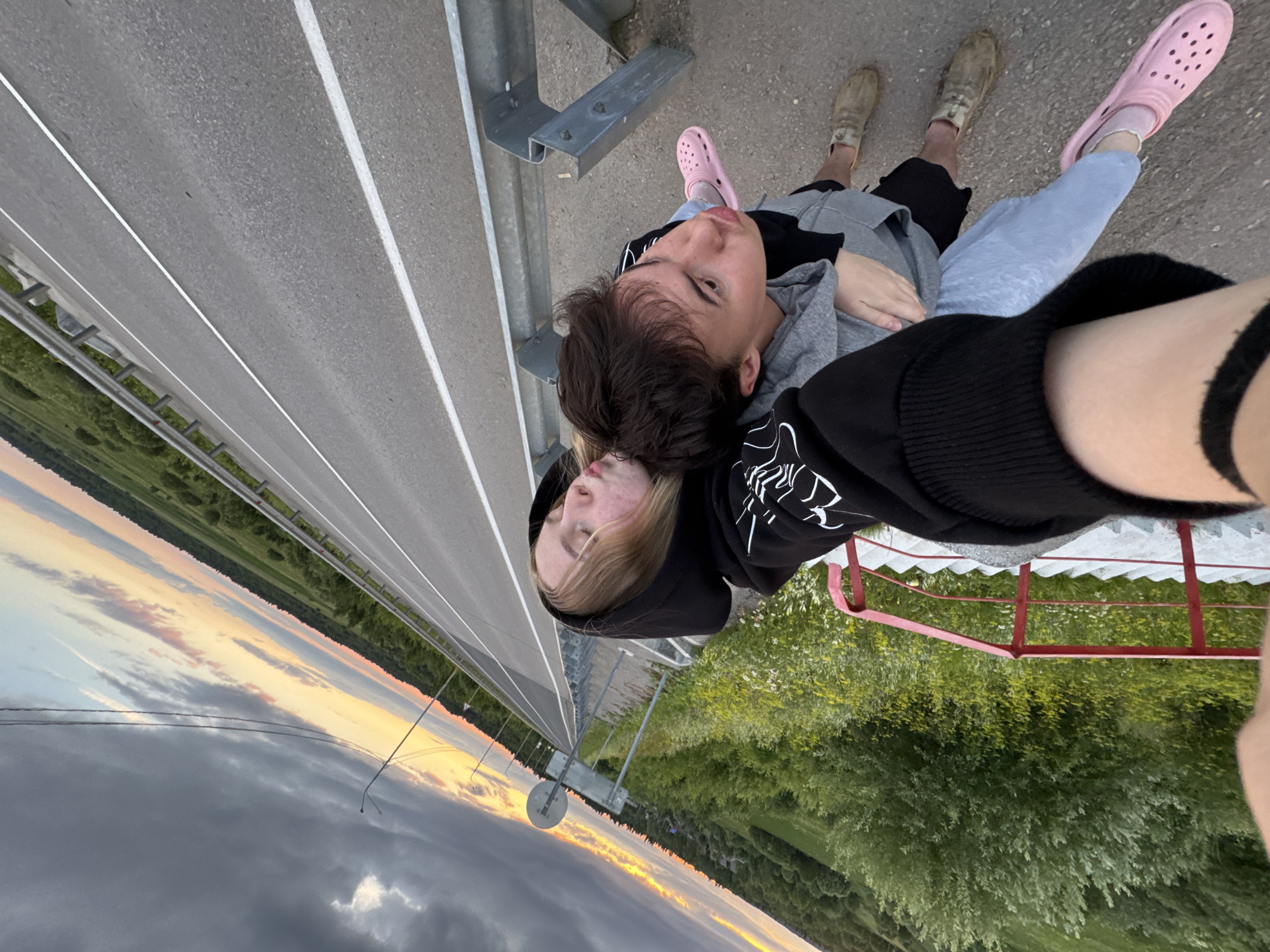 The image shows two young people embracing while taking a selfie on what appears to be a bridge or elevated walkway. The person in the foreground, on the right, is wearing a black hooded sweatshirt with white graphic text on the sleeve. Their arm is around the other person, who is leaning into them. This second person, on the left, is wearing a grey hooded sweatshirt and has their head resting on the first person's shoulder. They both have their eyes closed, suggesting a moment of peaceful intimacy. The background shows a road with white dividing lines, a metal guardrail, and a lush green landscape with trees. In the distance, the sky displays a colorful sunset with hues of orange, yellow, and pink mixing with dramatic dark clouds. The overall mood is serene and romantic, possibly captured during a quiet moment at dusk. The location is identified as Ryabinino, Russia.