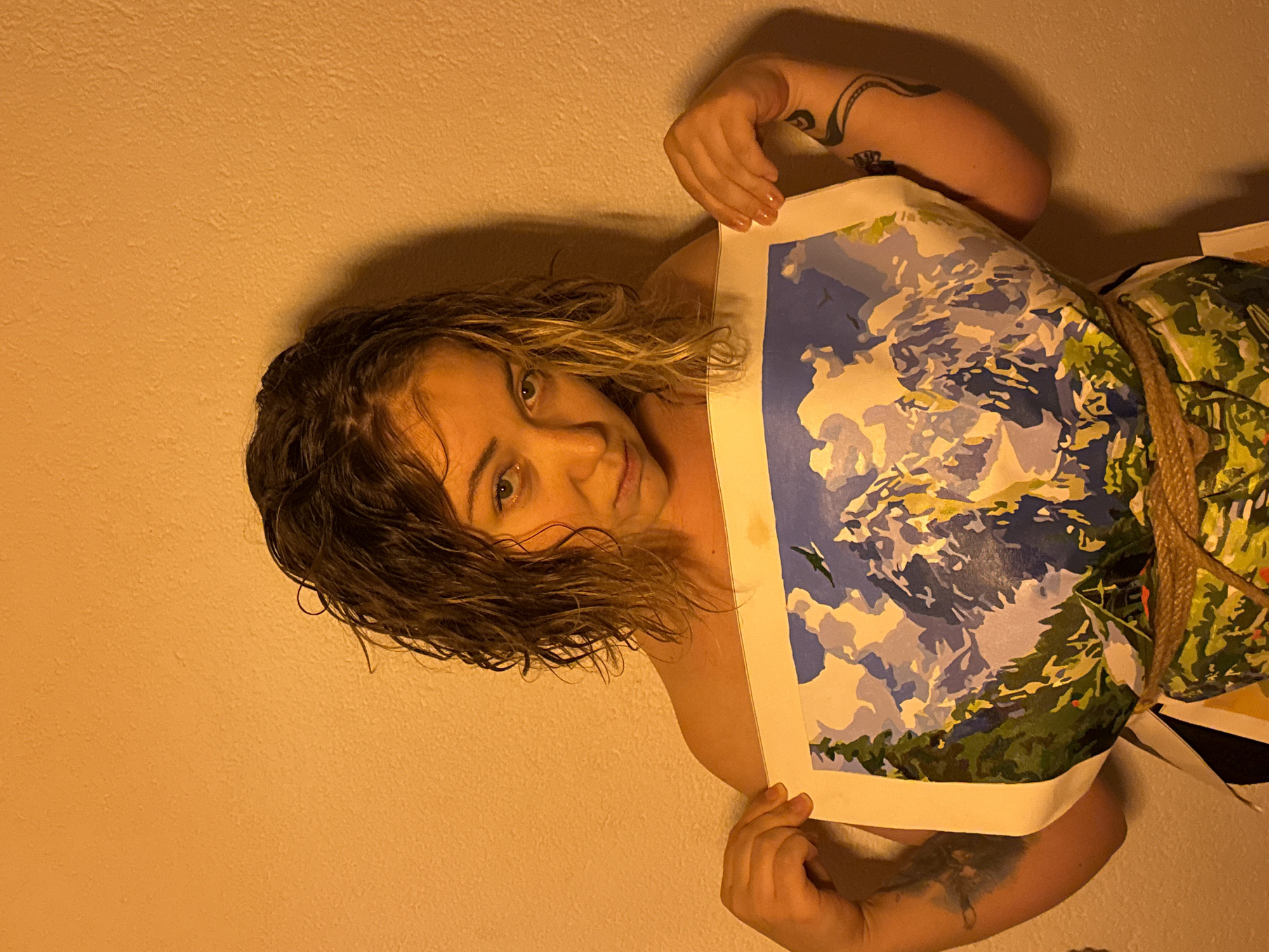 A woman with wet, blonde-brown hair is holding up a painting. The painting depicts a mountain range with blue and white sky. A rope is tied around her torso. She is standing in front of a light-colored wall. The location is unclear.
