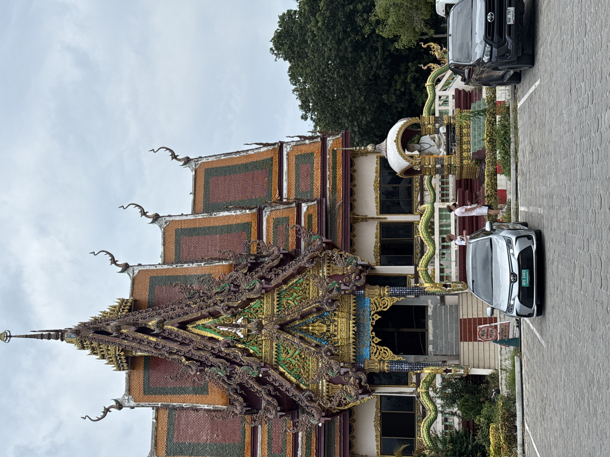 The image shows the exterior of a Buddhist temple in Ko Samui District, Thailand. The temple features an ornate, multi-tiered roof with colorful tiles and intricate golden decorations. In front of the temple is a parking lot paved with grey cobblestones. Two cars are parked: a silver sedan on the left and a black pickup truck on the right. A statue of a seated figure, possibly a monk, is visible in a shrine area to the right of the temple entrance. Several people are standing near the cars and the shrine. Lush green trees are visible behind the temple. The sky is overcast.