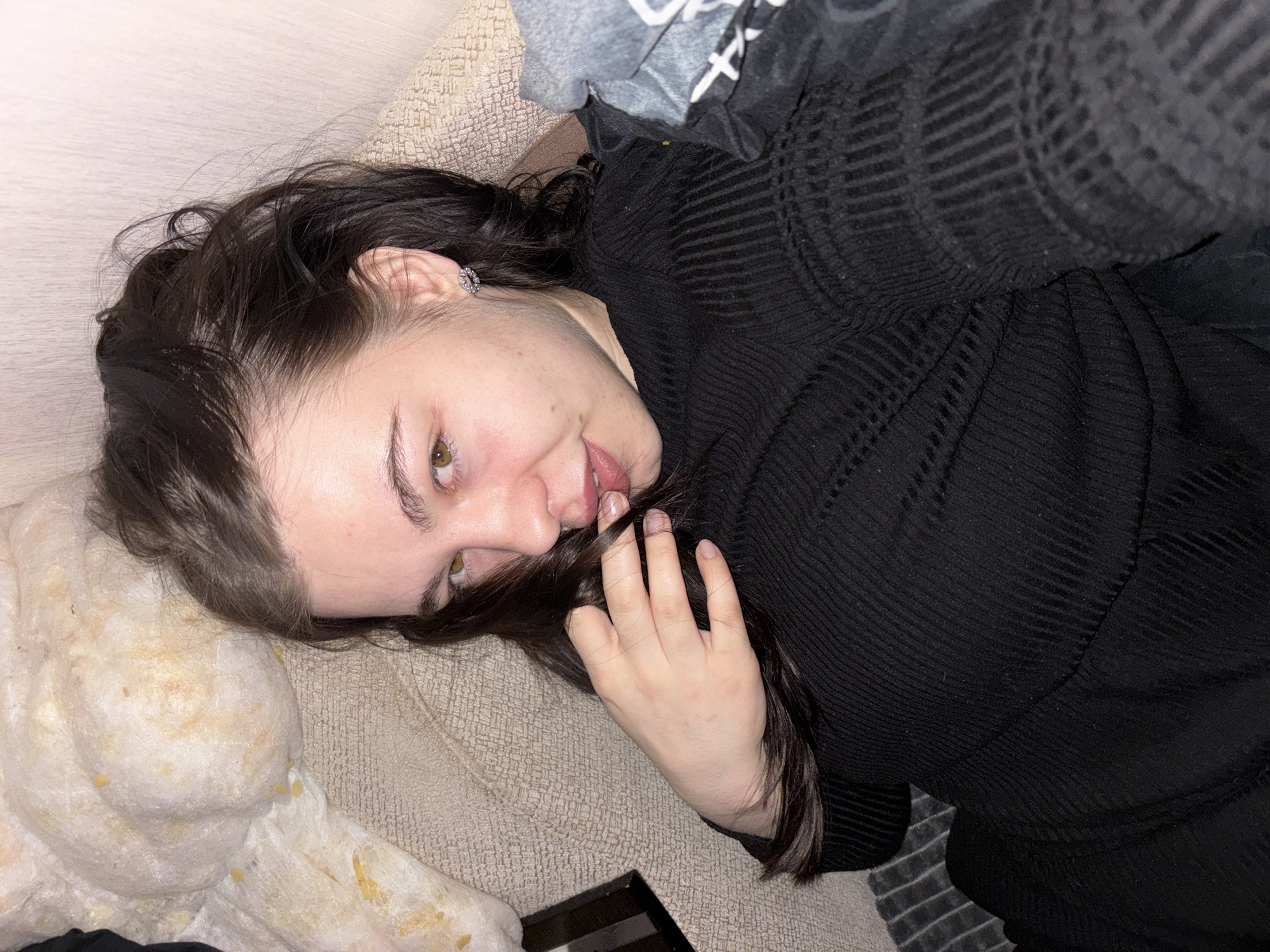 A woman with dark hair is lying on her back on a beige couch or cushion. She is wearing a black, long-sleeved top with a ribbed texture. Her head is tilted to the left, and her right hand is near her mouth, holding a strand of her hair. Her face is visible, showing a slightly open mouth. She has light skin and dark eyebrows. In the upper part of the image, there is a grey garment near the woman's shoulder. At the lower left of the image, there is a white, fibrous, and somewhat shredded material. The location of the image is unknown.