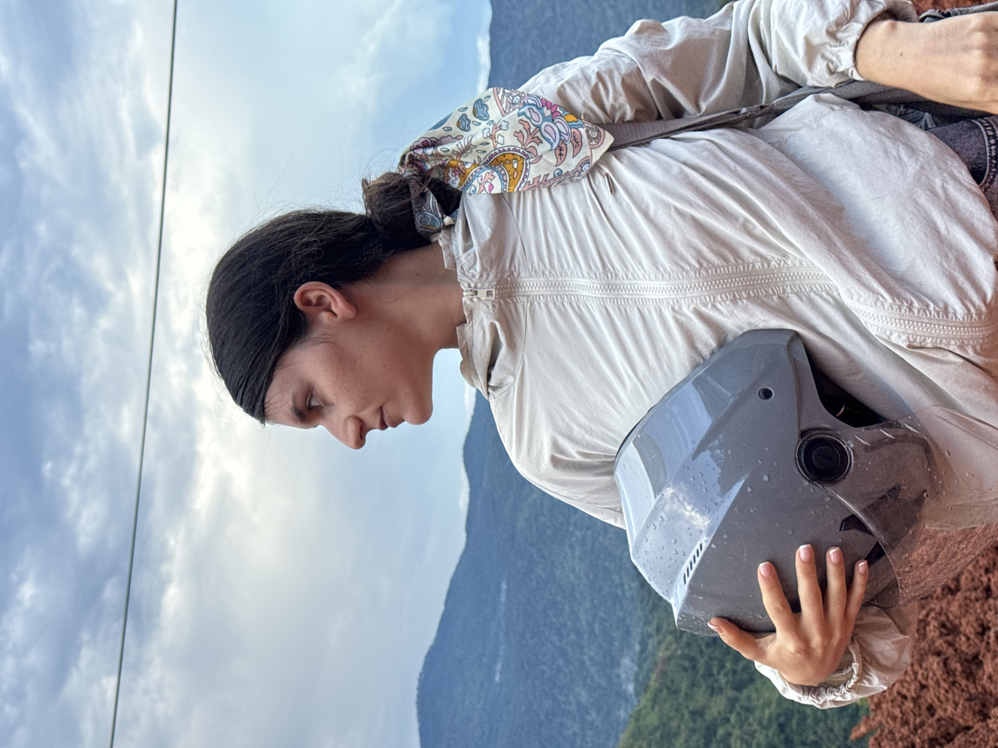 A young woman is captured in a medium shot, holding a grey motorcycle helmet in her left arm. She is wearing a light-colored, zippered jacket and a colorful patterned scarf tied around her ponytail. Her hair is dark and pulled back. She is looking off to her left, with a neutral expression. The background reveals a landscape of lush green mountains under a cloudy sky, hinting at a natural, possibly rural, setting. The woman's attire suggests she might be on an outdoor excursion. The ground in the foreground appears to be reddish-brown, possibly soil or clay. The image does not provide any specific landmarks or indications that can definitively pinpoint the location to the city of Nao Sẻ, Vietnam.