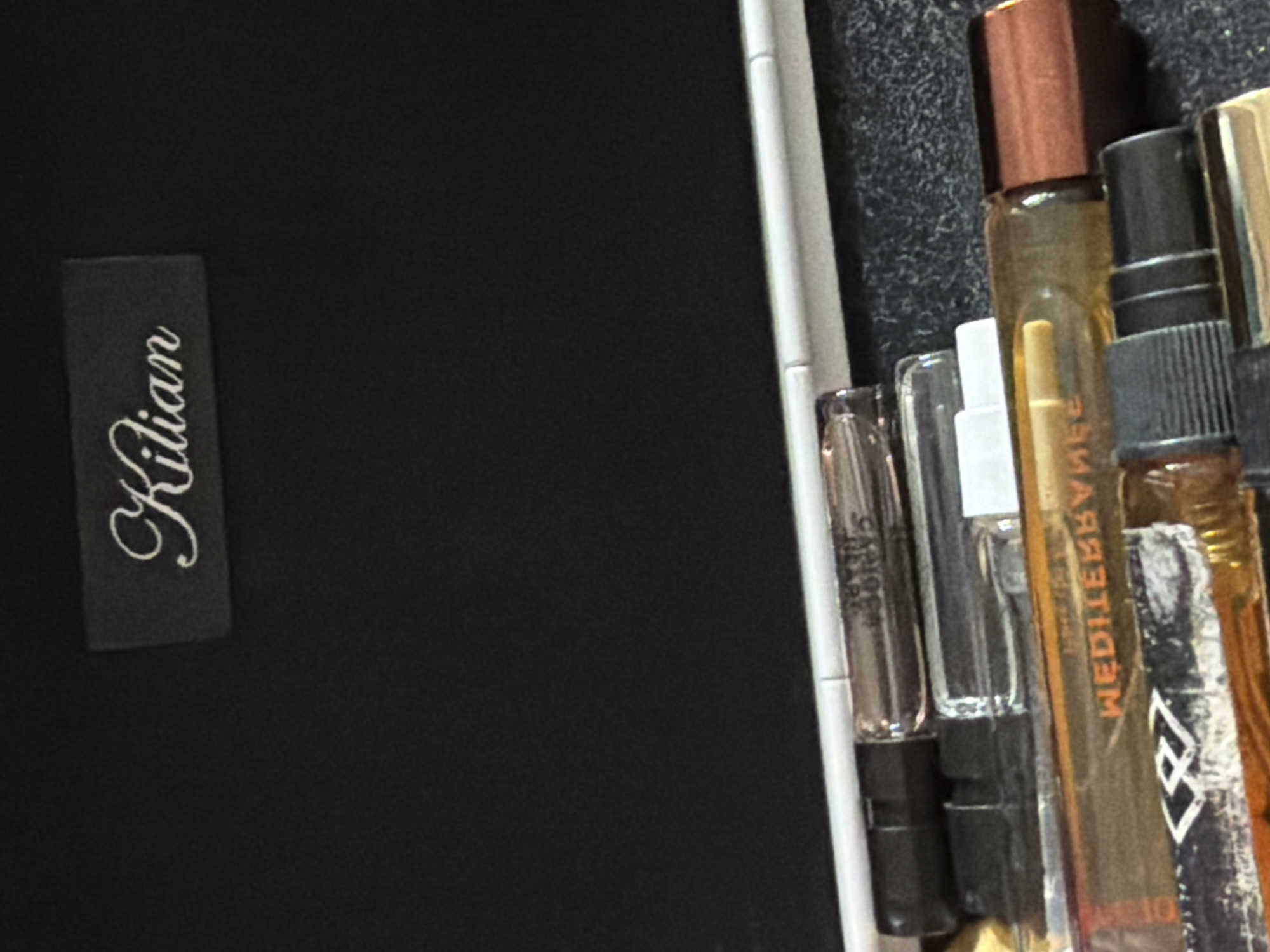 The image shows a black case with a "Kilian" label on the left side. The right side of the case contains several perfume sample vials of varying sizes and colors. The vials are arranged in a row. The background of the inside of the case appears to be a dark, textured material.