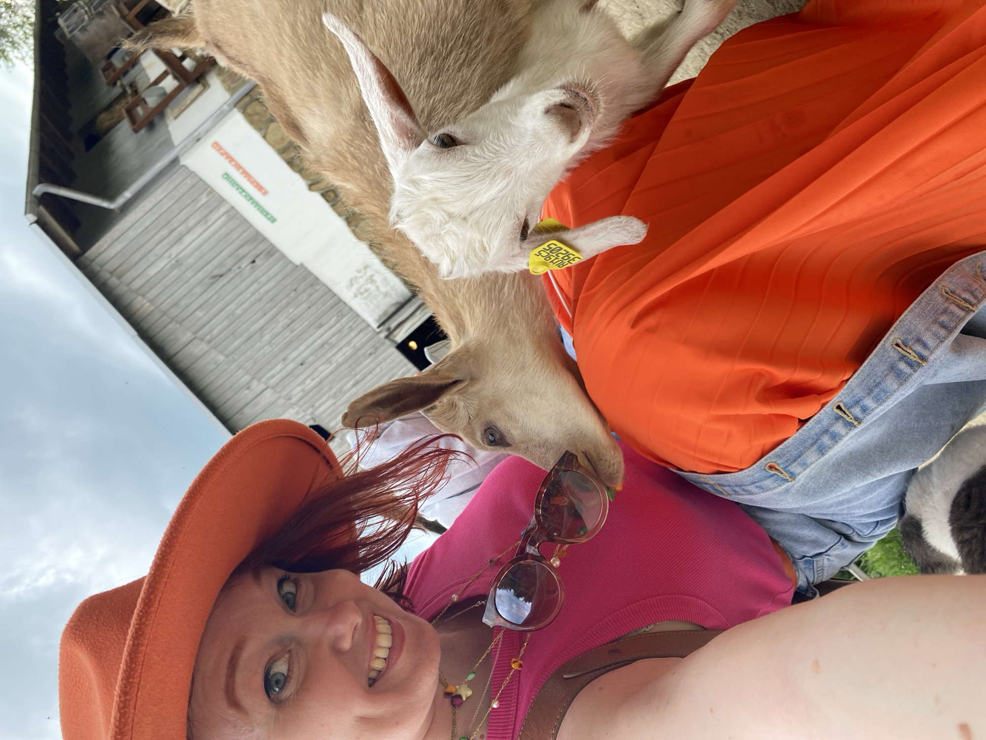 A selfie shows a woman with red hair, wearing an orange hat, pink shirt, a necklace with colorful beads, and sunglasses, smiling. Two goats are interacting with her, one light brown and the other white with a yellow tag in its ear. The woman is wearing a denim jacket over an orange shirt. The background includes a building with text that appears to say "ЕЗИММАЧЕРКАВЛО." The location is possibly Прогресс, Russia, based on the text.