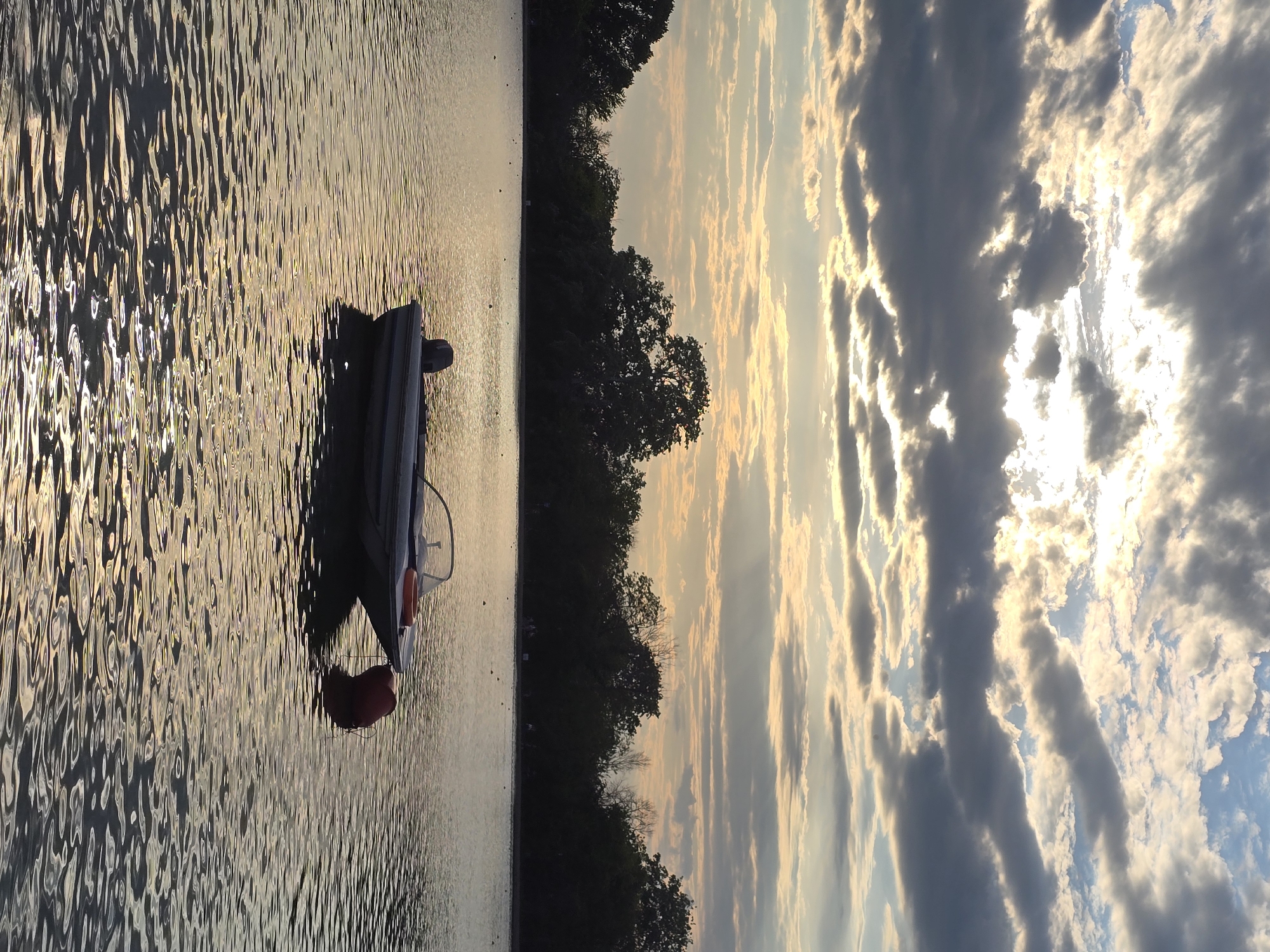 The image shows a lake with a small boat floating on the water. The sun is setting, casting a golden light on the water and the clouds in the sky. The location is Unknown, Unknown. The scene consists of water, sky, trees, and a boat with an orange float.