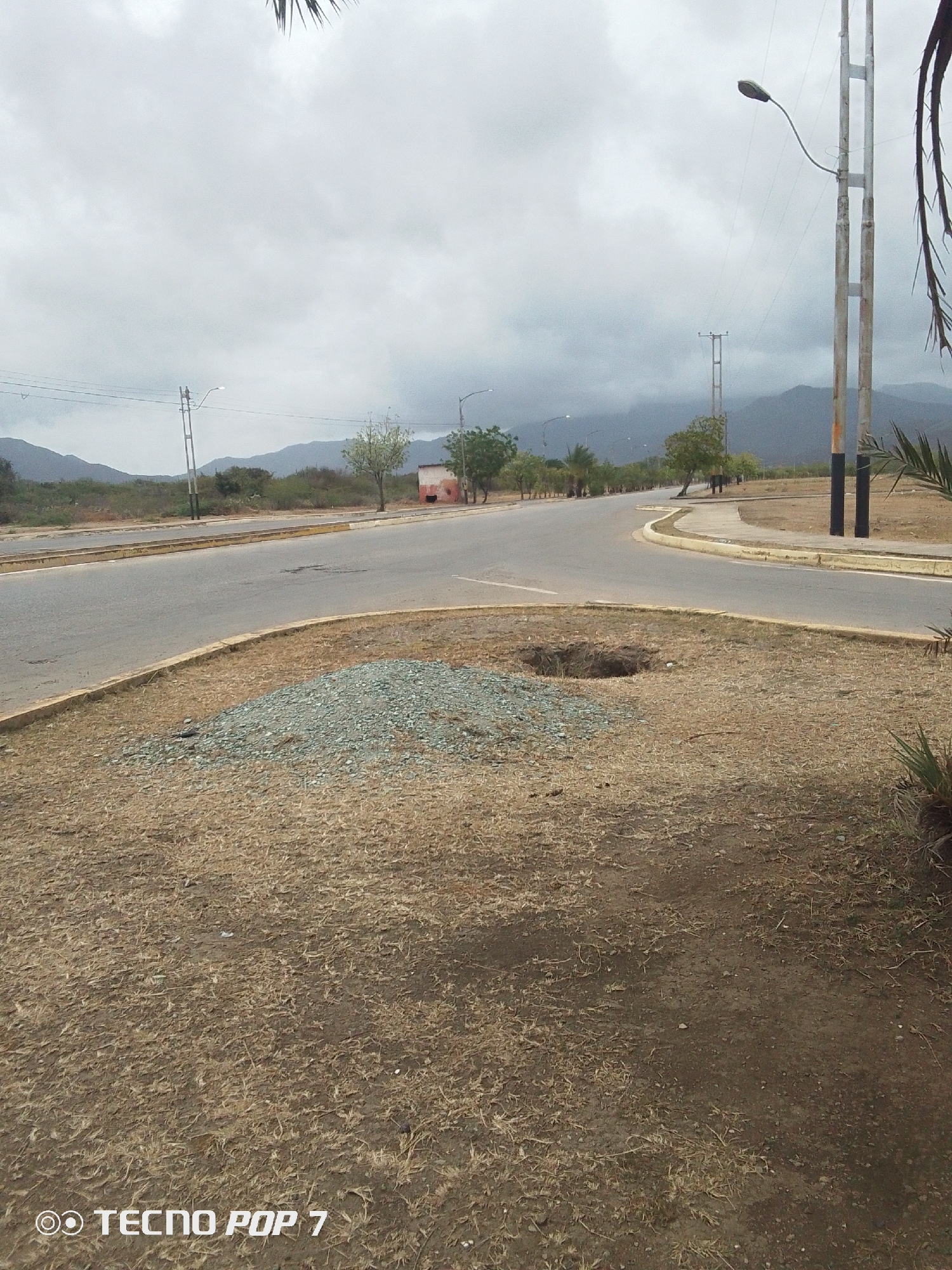The image captures an outdoor landscape in Altagracia, Venezuela, depicting a road junction under an overcast sky. In the foreground, the ground is covered with dry, sparse brown grass and bare earth. A small pile of light gray gravel sits on the left side of the foreground, next to a dark, circular hole dug into the ground.
Two paved roads diverge from the viewer. The road to the left is wider and extends into the distance, while the road to the right curves around a grassy median. Both roads are bordered by concrete curbs with yellow painted edges. Several tall, metal utility poles with streetlights are positioned along the roads, some featuring black and white painted sections at their bases.
The middle ground shows more dry vegetation, including scattered trees and shrubs with light green foliage, indicating sparse plant life. A small, possibly red or pink building with a dark roof is visible in the distance along the left road.
In the background, a range of dark mountains stretches across the horizon, appearing somewhat hazy or muted due to the atmospheric conditions. The sky is uniformly covered with light gray clouds, suggesting an overcast day with diffuse lighting. There are no people or vehicles visible, creating a quiet and somewhat still atmosphere.
The bottom-left corner of the image displays text that reads "OO TECNO POP 7", indicating the device used to capture the photograph.