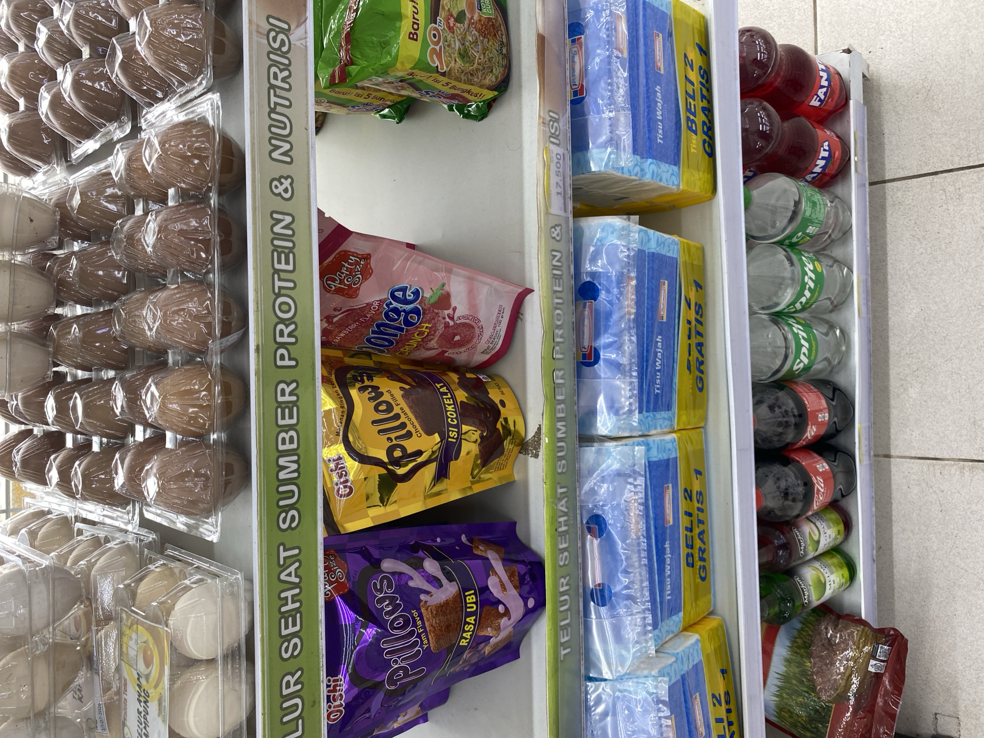The image shows a store shelf stocked with various grocery items. The top shelf displays rows of eggs in plastic containers. Below that, the middle shelf holds bags of "Pillows" snacks, one purple and one yellow, along with a pink bag of "Bonge" snack. To the right, a green bag of instant noodles is visible. The shelf below that is filled with packages of "Tisu Wajah" (facial tissues) with a "Beli 2 Gratis 1" (Buy 2 Get 1 Free) promotion. The bottom shelf contains several bottles of drinks, including Sprite and Fanta. A red bag of what appears to be rice is positioned on the far left of the bottom shelf. The overall environment suggests a retail setting, likely a convenience store or a small supermarket. The lighting indicates indoor illumination. No people are visible in the image.