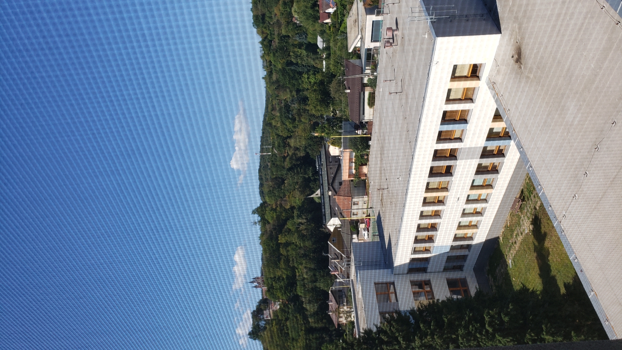 The image depicts a view from a window overlooking a cityscape with a clear blue sky. In the foreground, there is a white, multi-story building with a flat roof. This building has numerous windows with wooden frames, some of which are open. A portion of another building, also with a flat roof, is visible in the lower right of the image.
Beyond the foreground building, a small courtyard or grassy area is visible, featuring a solitary bench. Further in the background, a collection of residential houses with red-tiled roofs are situated on a gentle slope. Behind the houses, a dense green forest covers the rolling hills. On one of the hilltops, a distinctive red building with towers, likely a church or historical monument, can be seen. In the distance, a tall communication tower rises above the trees on another hill.
The scene appears to be a typical urban or suburban landscape under a bright, sunny sky, suggesting a daytime setting. The presence of the window screen in the foreground indicates the photograph was taken from indoors. The architectural style of the buildings suggests a Central European context, possibly Prešov, Slovakia, given the mention in the prompt. However, without specific identifying landmarks or context beyond what is visually evident, a definitive location within Prešov cannot be confirmed.