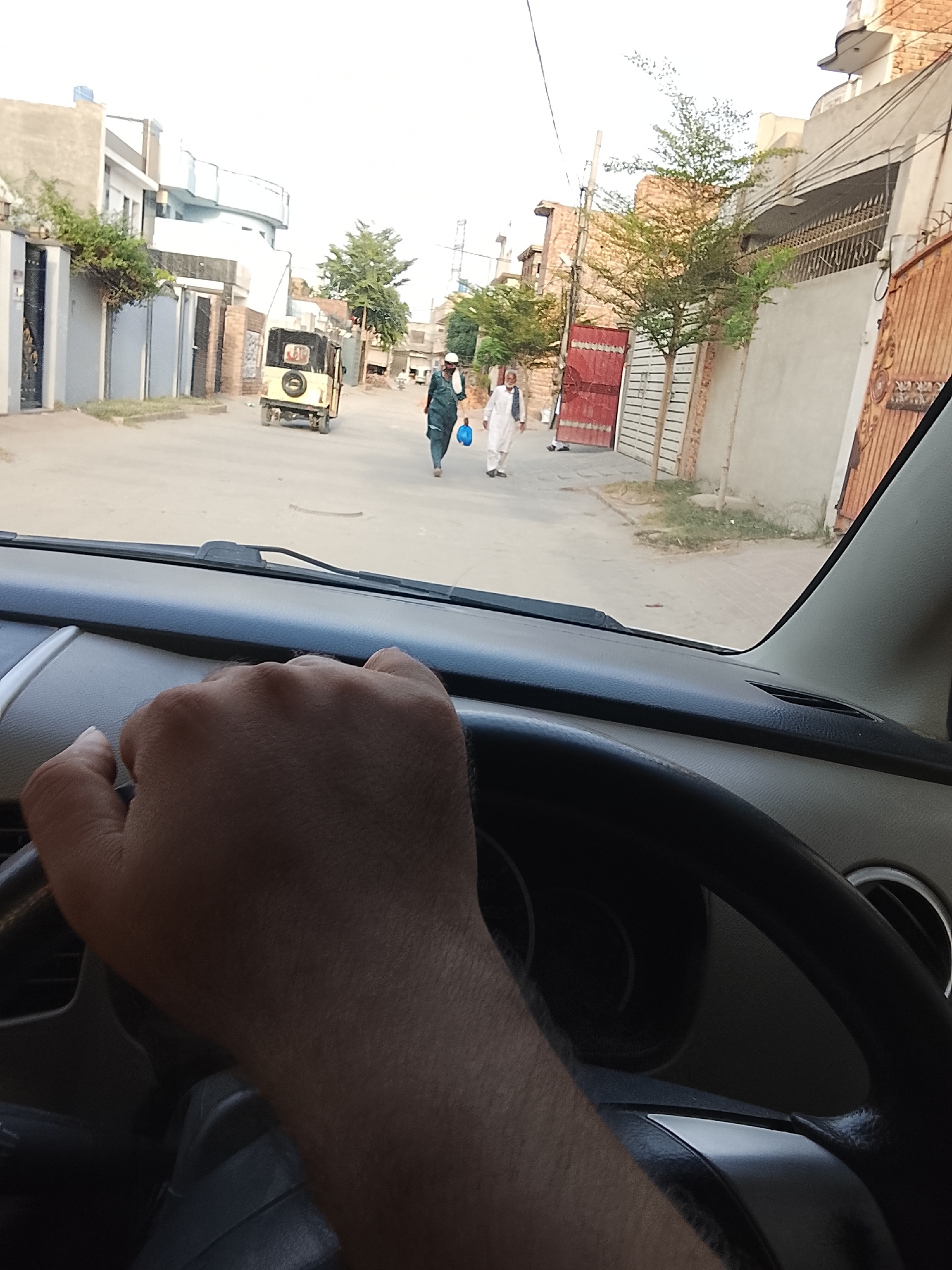 The image shows a view from inside a car, presumably the driver's perspective. A hand is visible on the steering wheel. Through the windshield, a street scene in Nasheman Colony, Pakistan, is visible. Two men are walking on the street; one is wearing a white thobe, and the other is wearing a dark robe while carrying a blue plastic bag. A yellow auto-rickshaw is parked in the distance. Buildings line both sides of the street; some have gated entrances. Trees are planted on the sides of the road. Overhead power lines are visible. The sky is overcast.