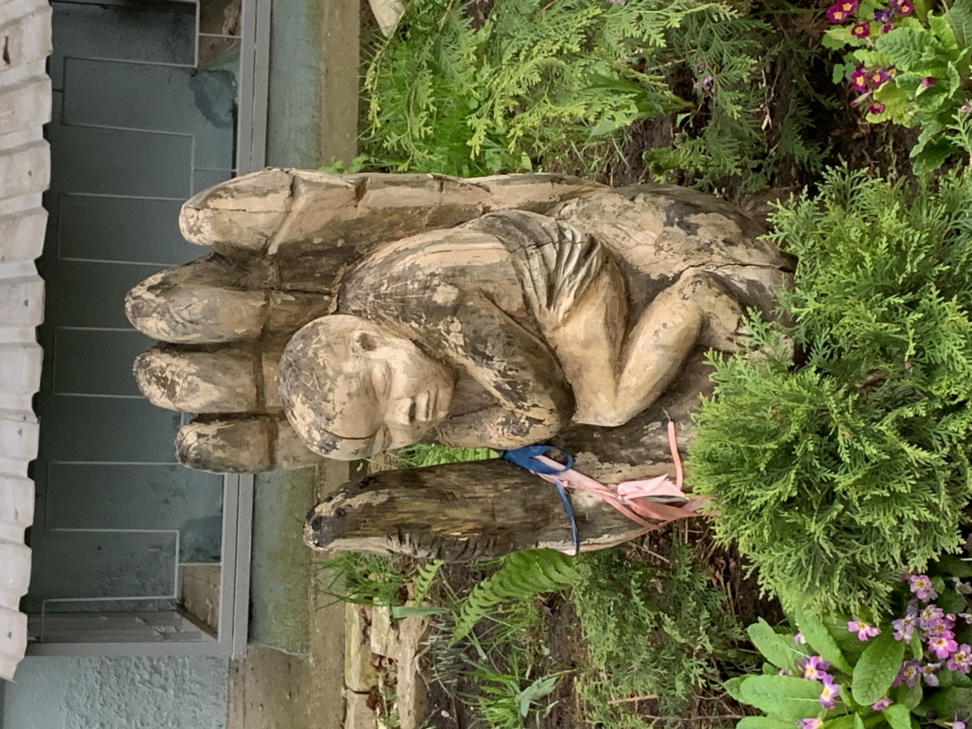 The image depicts a wooden sculpture of a human figure nestled within a large hand. The sculpture is located outdoors, surrounded by green plants and flowers. The environment appears to be a garden or yard. A building with a blue-green facade is visible in the background, suggesting the sculpture is located near a structure. The image does not provide enough information to confirm it is in the city of Chaikovsky, Russia.