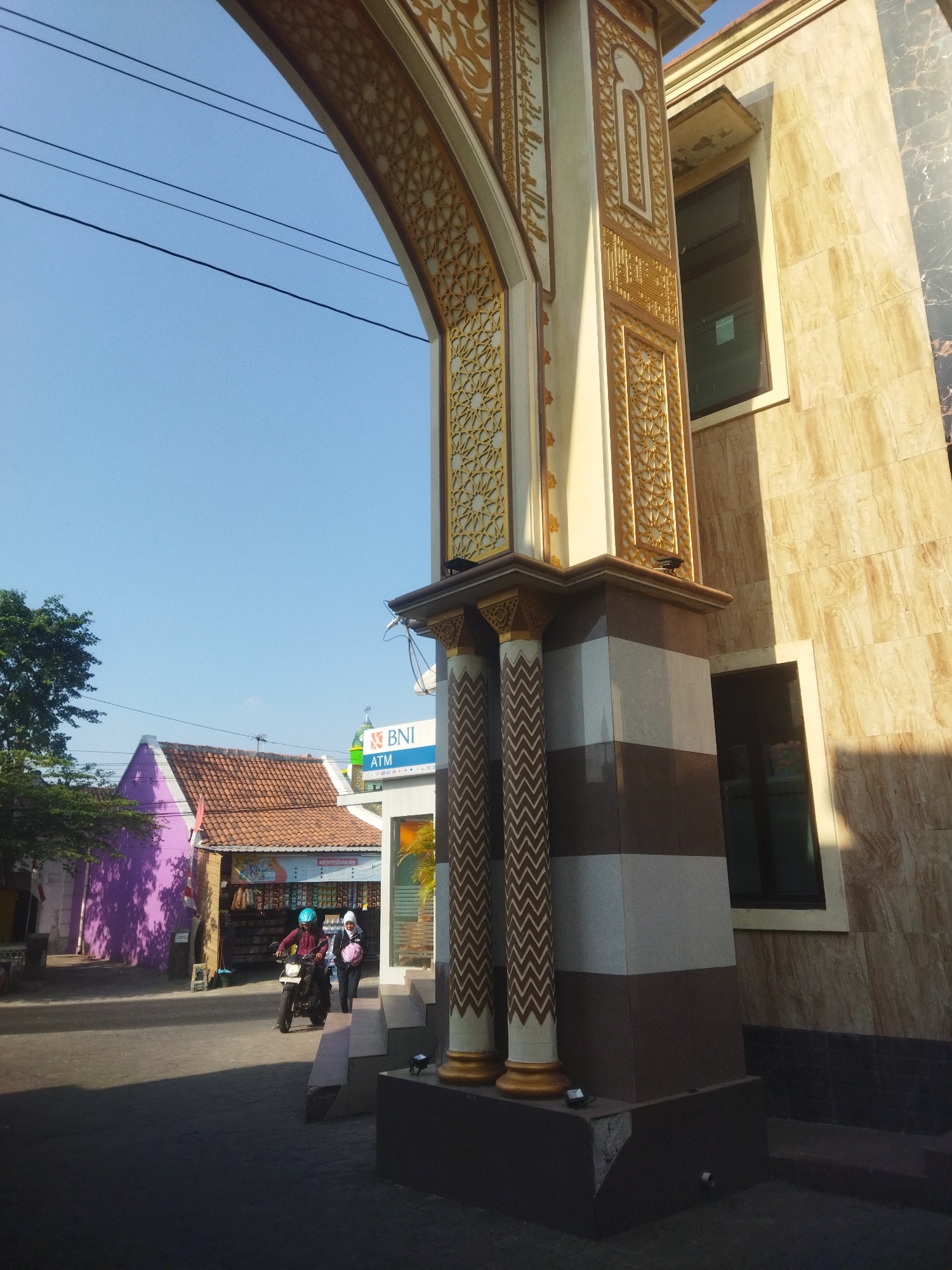 The image, taken in the city of Juwiring, Indonesia, captures a street scene dominated by an ornate archway and a building with a beige marble facade. The archway, with its intricate gold geometric patterns and Arabic calligraphy, frames a clear blue sky. Below the arch, two pillars with chevron patterns stand prominently.
On the left side of the image, a vibrant purple house with a terracotta-tiled roof is visible. Next to it, a row of shops with awnings lines the street. A person wearing a blue helmet rides a motorcycle, heading towards the archway. Another person, wearing a white hijab and carrying a pink bag, walks behind them.
In the background, a BNI ATM sign is attached to a building, and a green dome of what appears to be a mosque is visible above the shops. The sunlight casts long shadows across the paved street, indicating it is likely morning or late afternoon. The overall impression is a blend of traditional Indonesian architecture and modern urban elements.